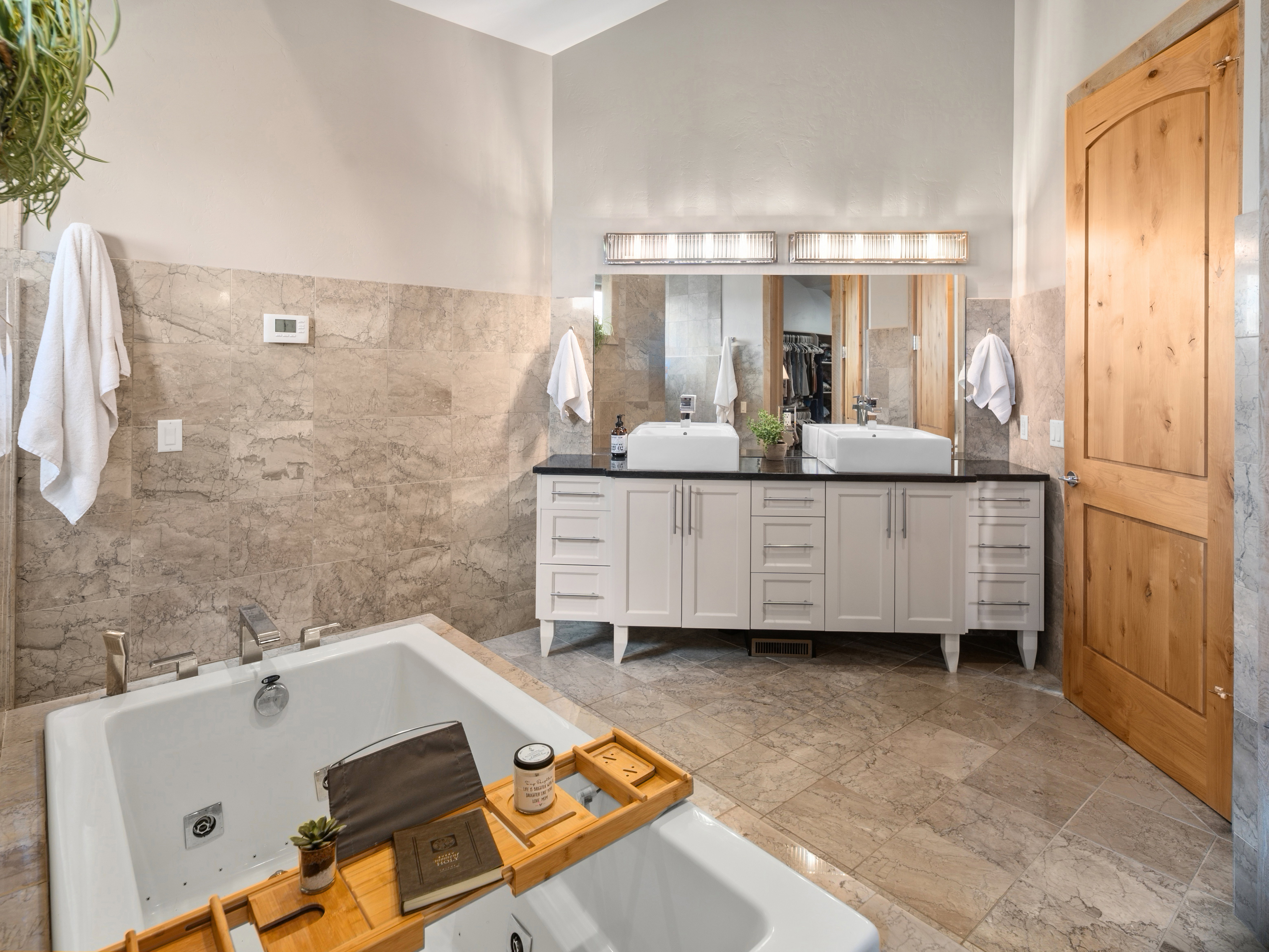 Built-in soaking tub with deck-mounted fixtures, dual vanity with vessel sinks, black countertops, and light cabinetry