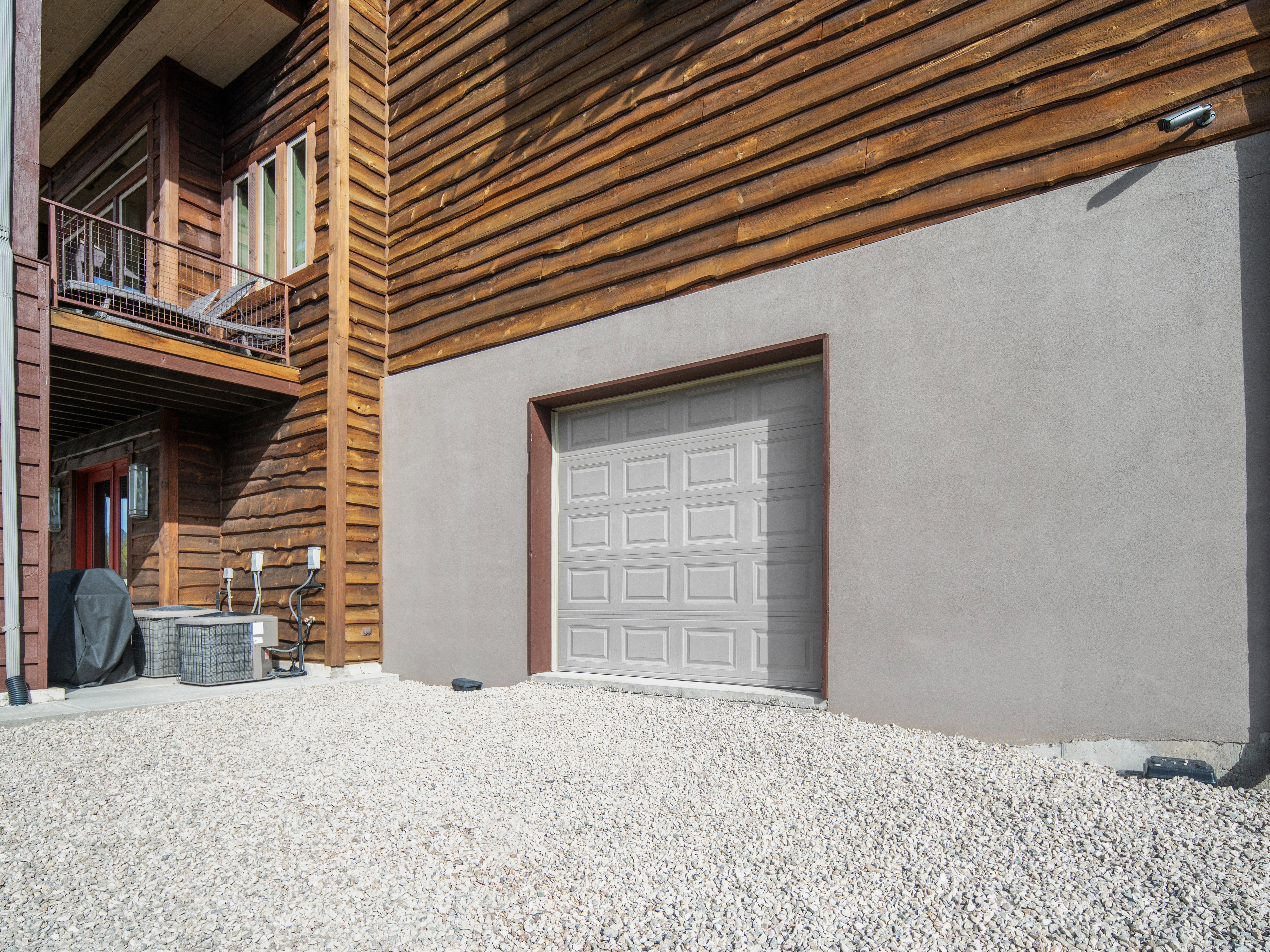 Stucco and wood-paneled exterior featuring a panel-style garage door and a gravel driveway