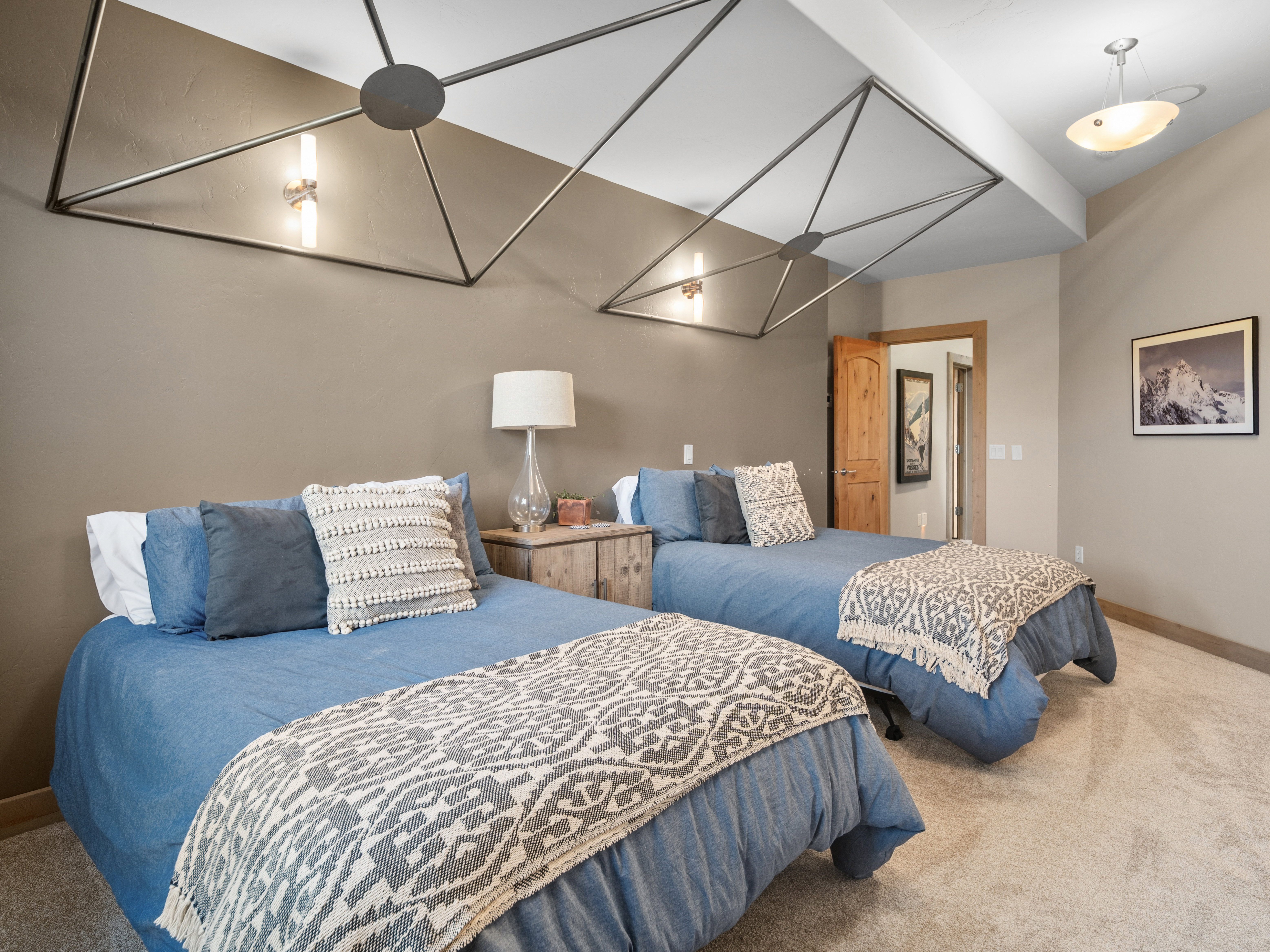 Two beds featuring neutral-toned carpeting, a light wood-finish nightstand, a wood-finish door frame, and geometric ceiling light fixtures