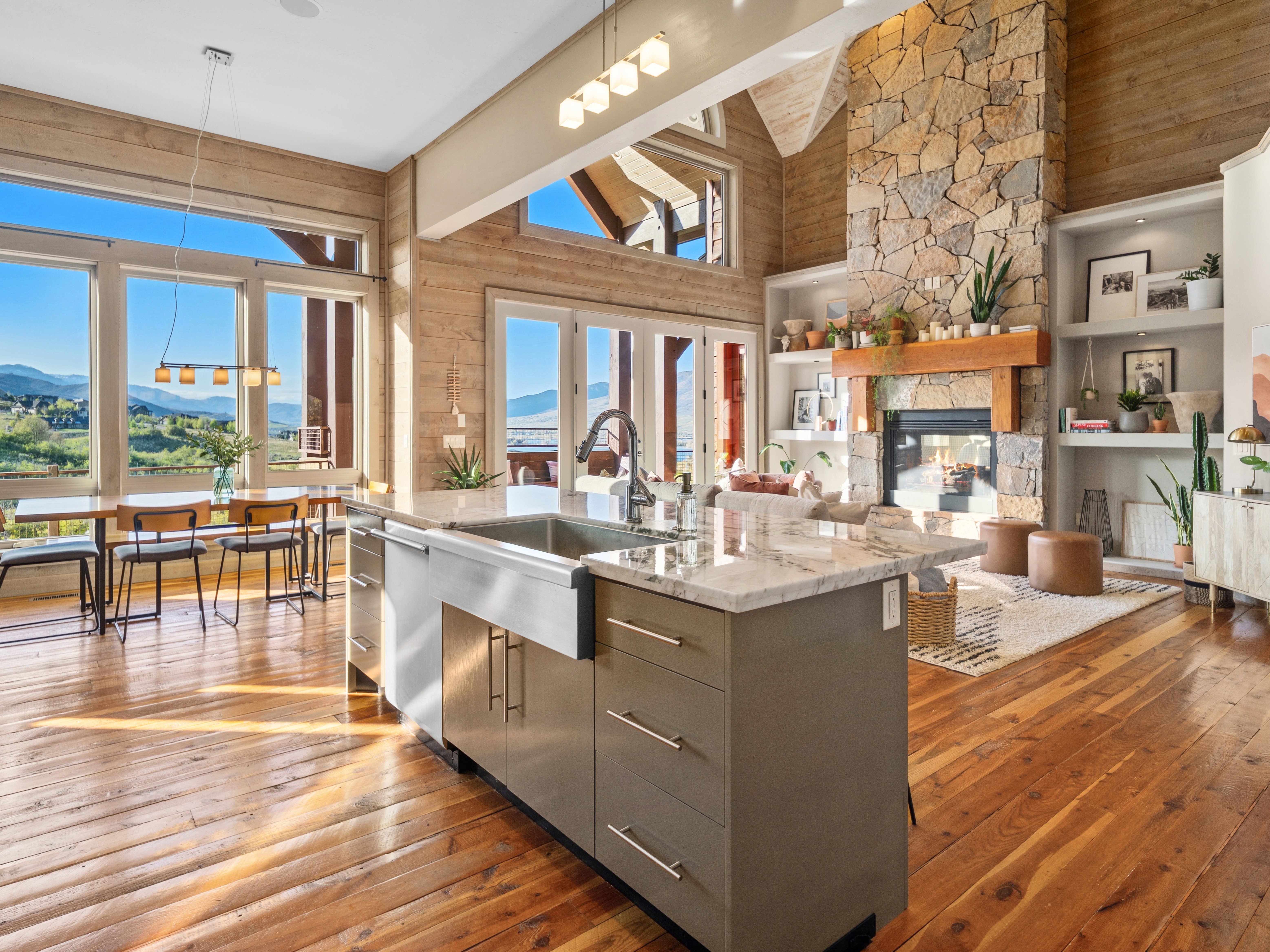Open-concept living space featuring expansive windows, wood-finish flooring, a stone fireplace with a substantial wood mantle, a kitchen island with a farmhouse sink, and integrated shelving units
