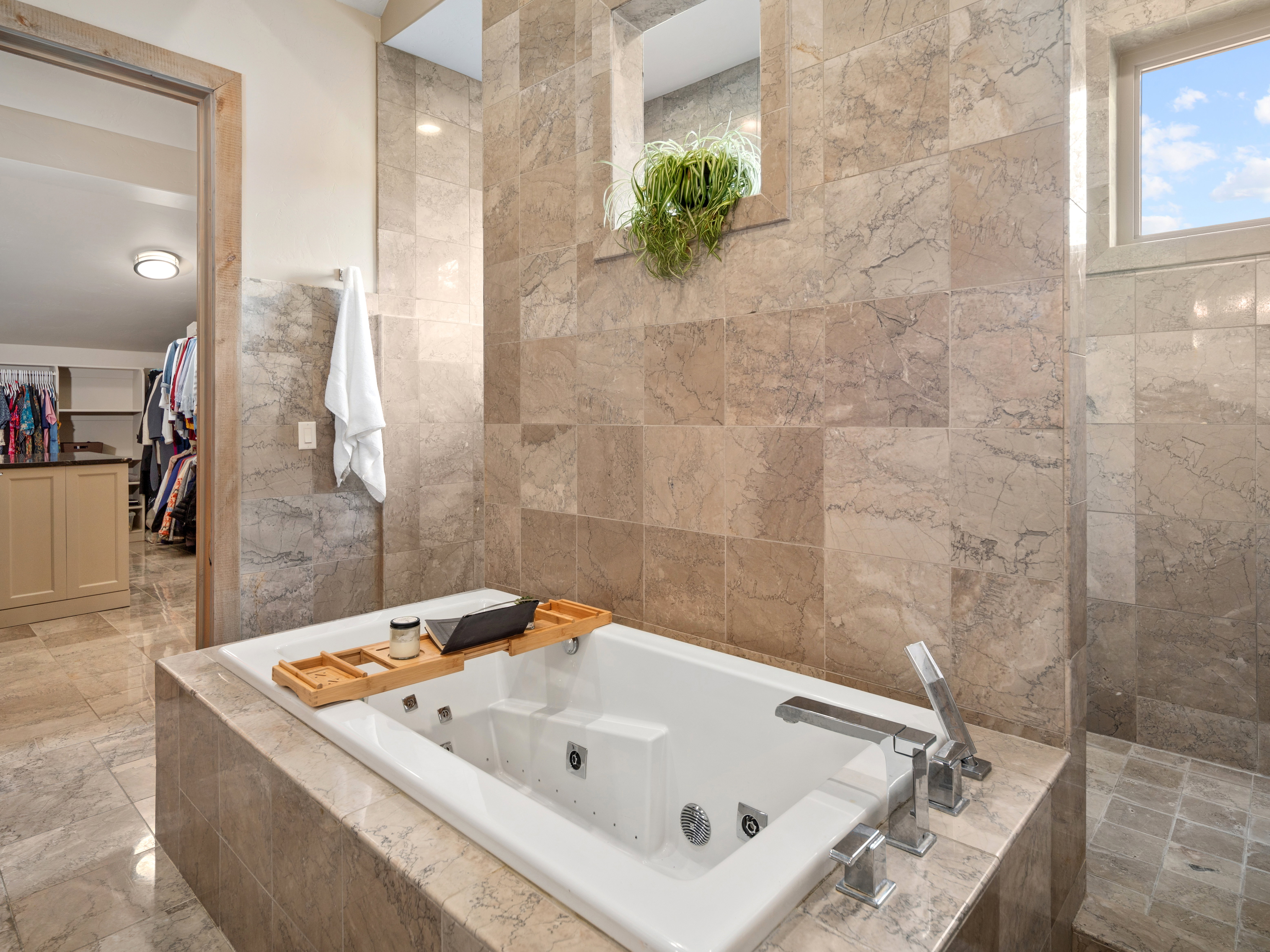White soaking tub with deck-mounted chrome fixtures, surrounded by large format natural stone wall and floor tiles