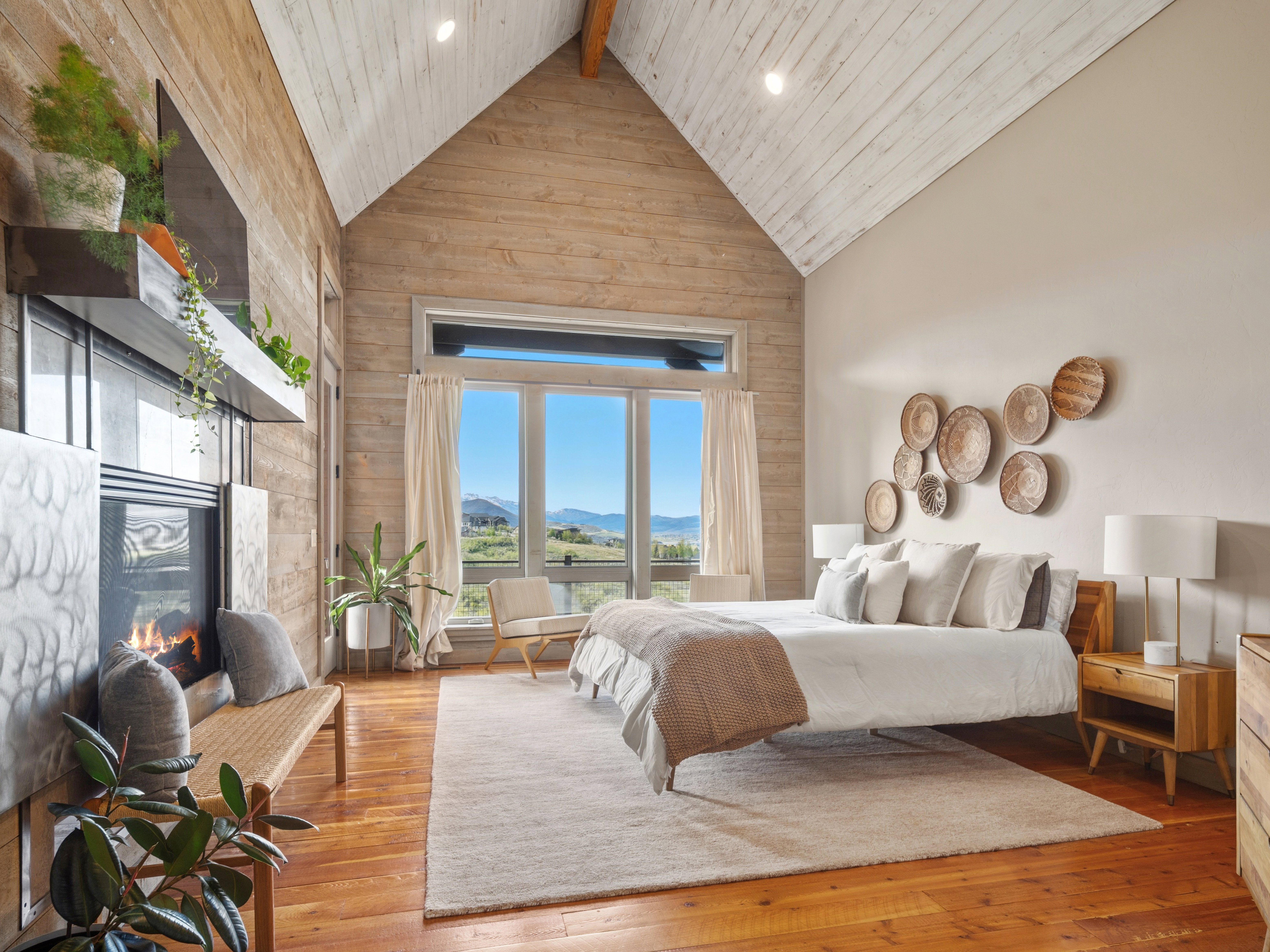 Vaulted ceiling with exposed beam, wood-finish flooring, full-height window with mountain scenery, wall-mounted fireplace, and light wood plank accent wall