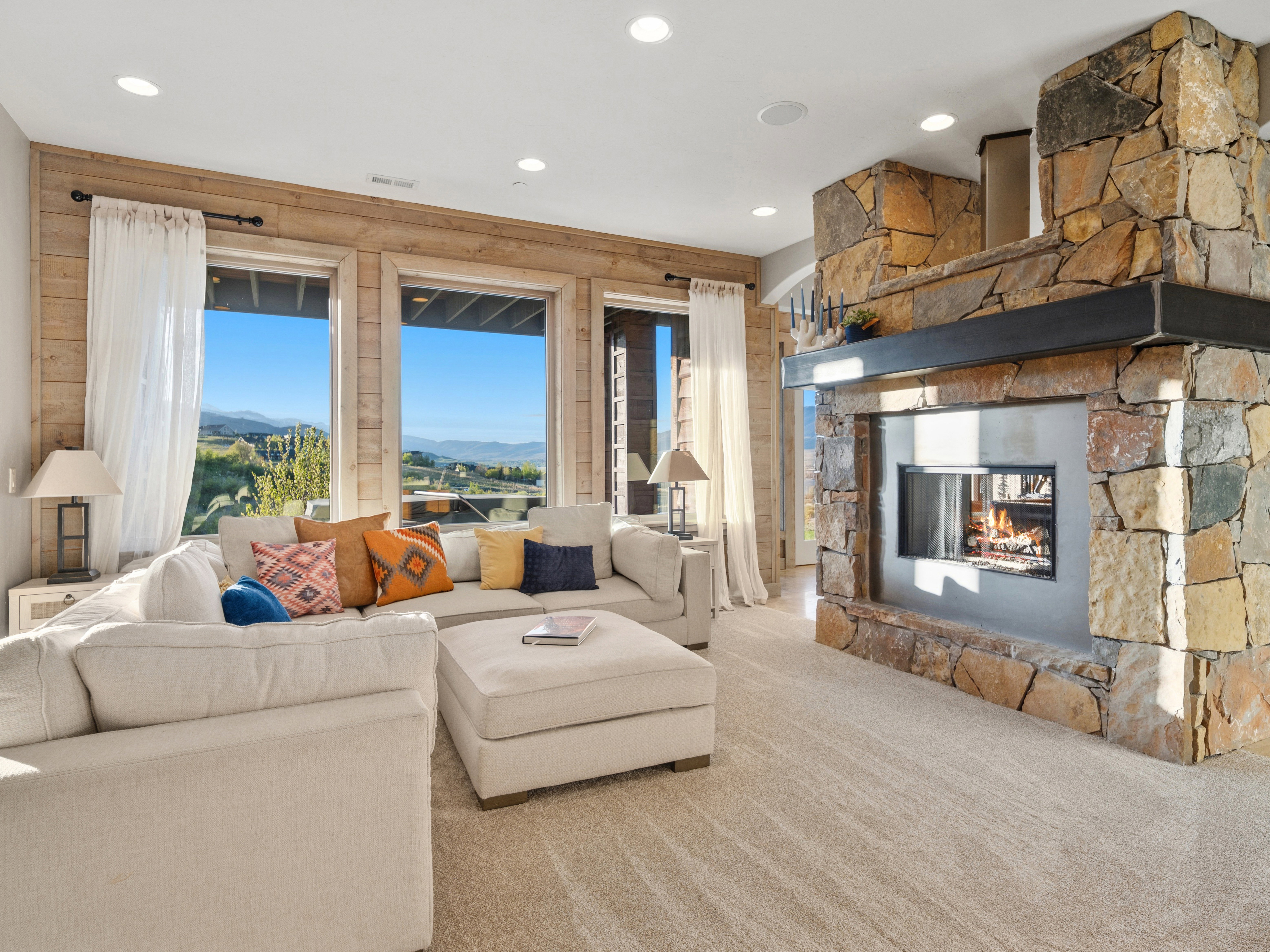 Spacious living area featuring a stone-clad fireplace with a metal mantel, recessed lighting, and large windows with surrounding wood paneling