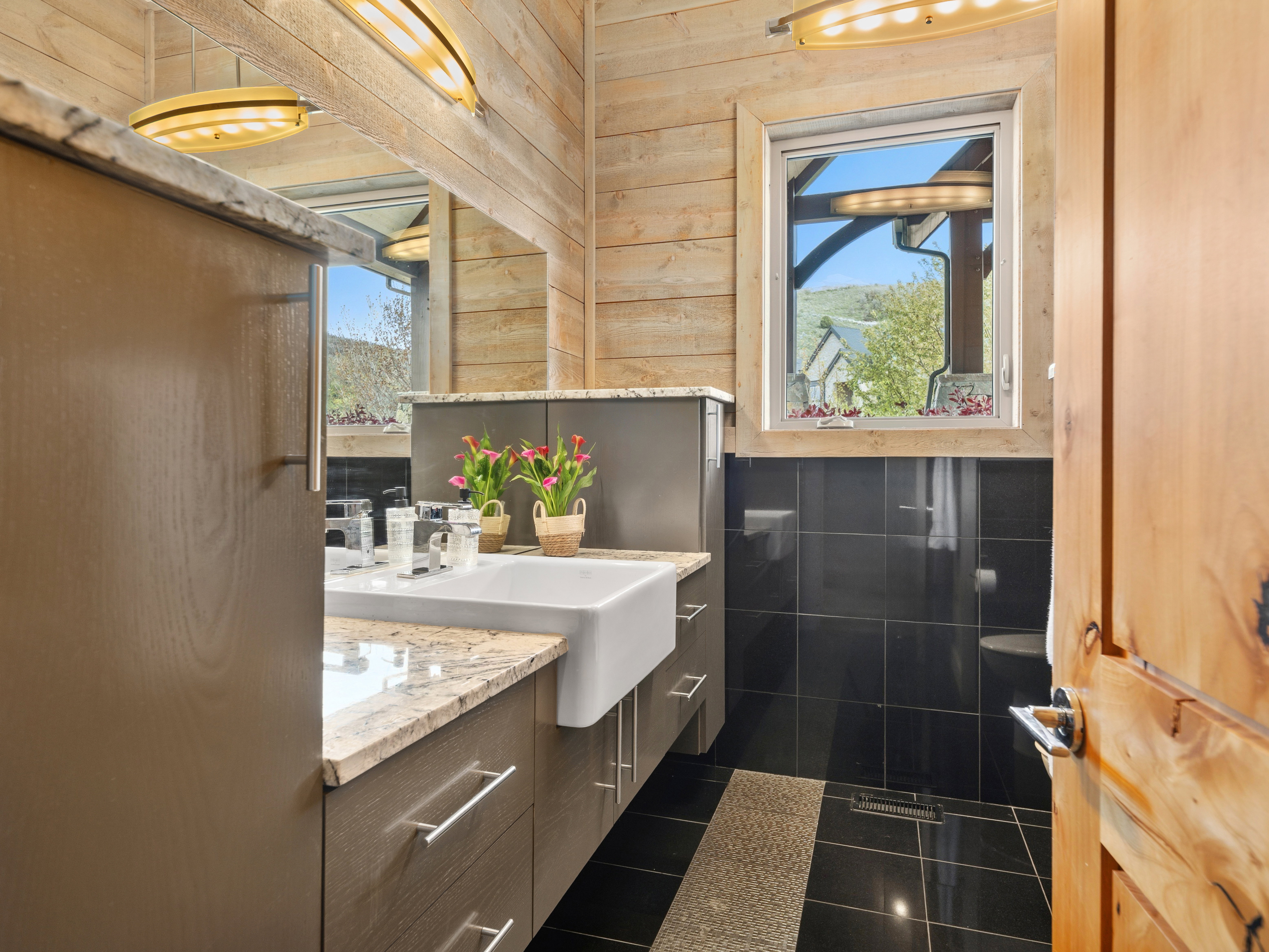 Bathroom featuring wood-paneled walls, black tile flooring with a stone accent, a window with a white frame, a vanity with a stone countertop, and a white farmhouse-style sink