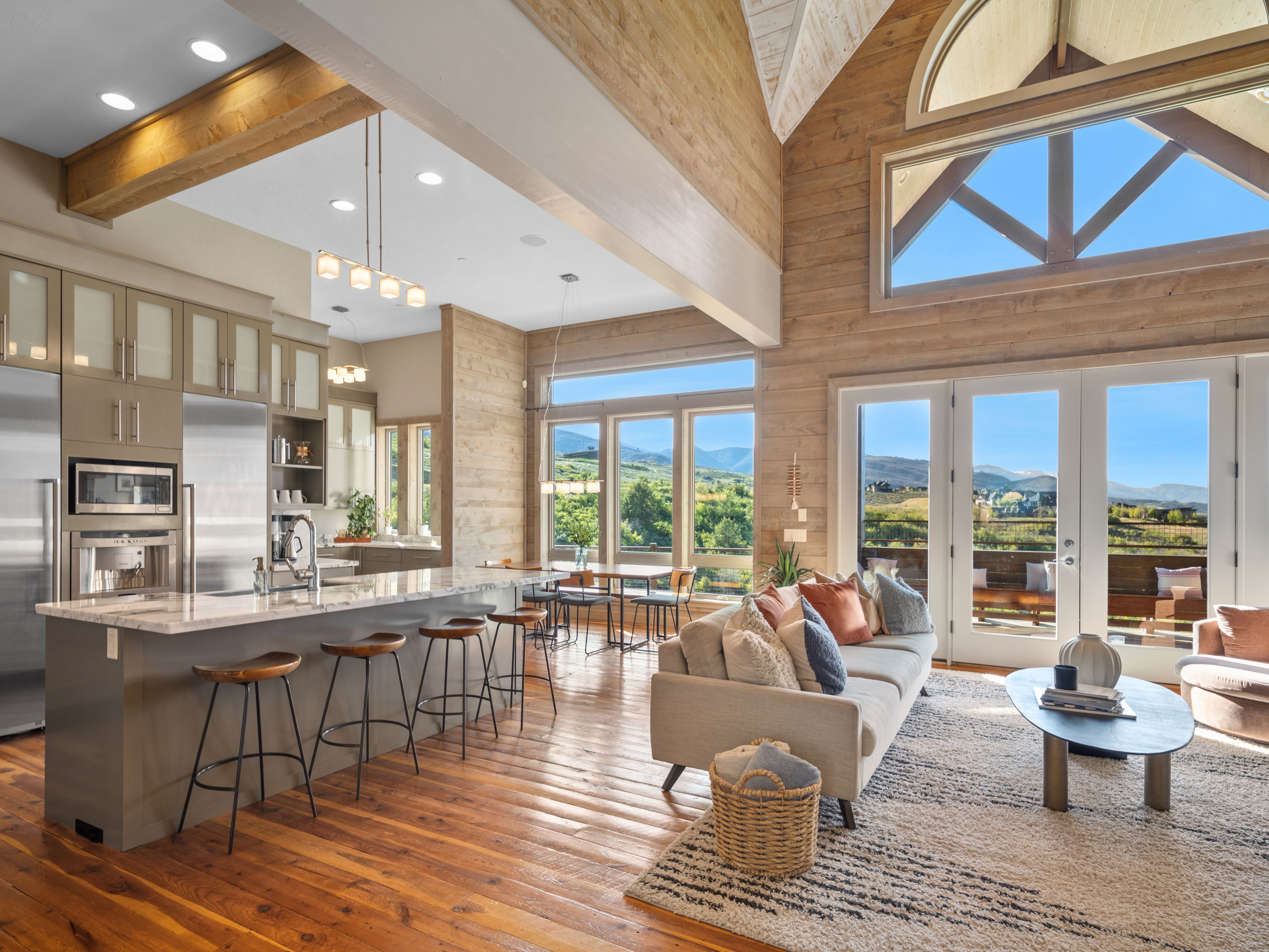 Open-concept living area featuring a vaulted ceiling with exposed beams, wood-finish flooring, a kitchen island with stone countertop, stainless steel appliances, and French doors opening to an outdoor area with mountain scenery