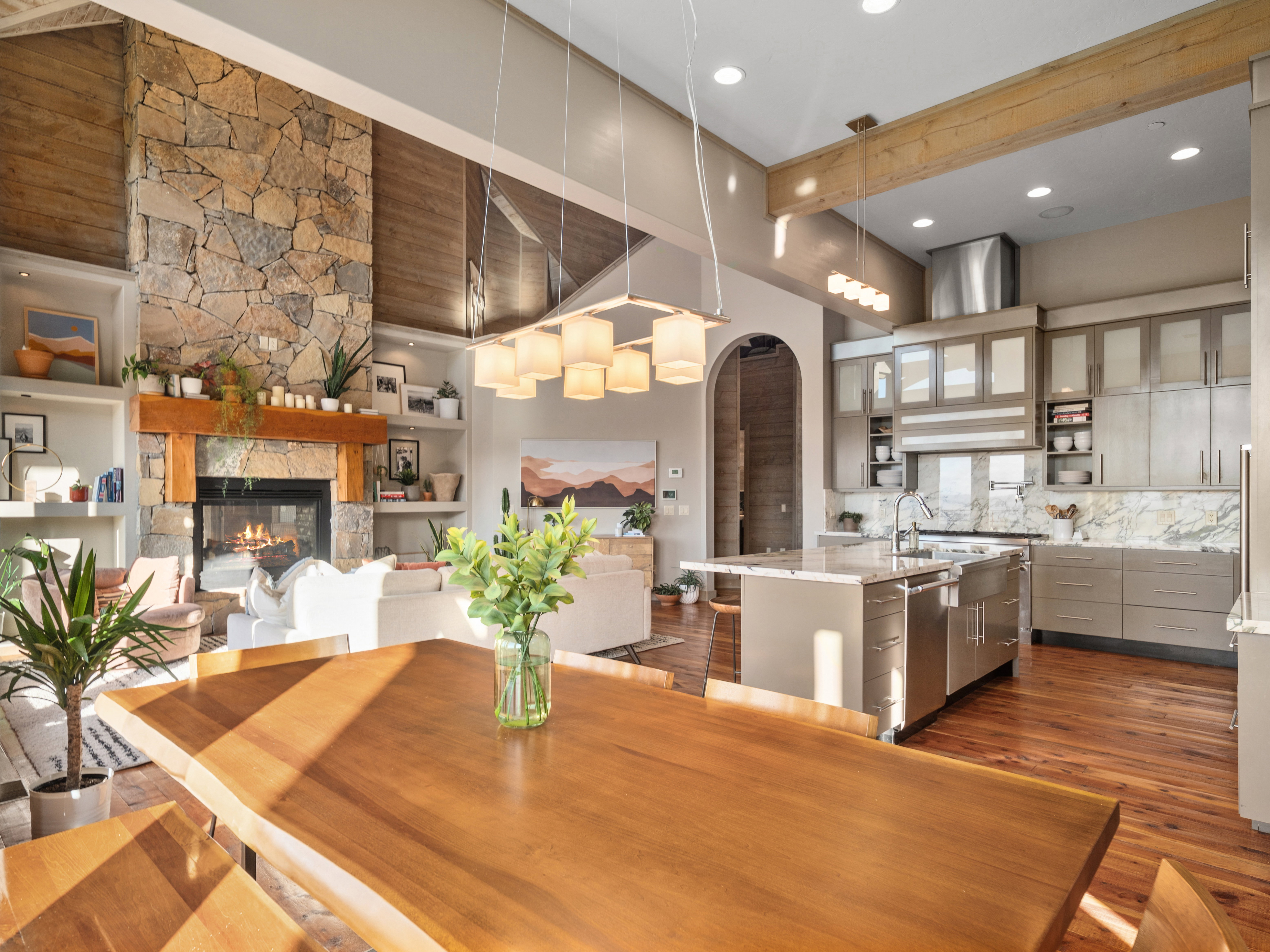 Open-concept living space featuring a substantial wood-finish dining table, a floor-to-ceiling stone fireplace with a substantial timber mantel, and a kitchen with an island, stone backsplash, and stainless-steel appliances