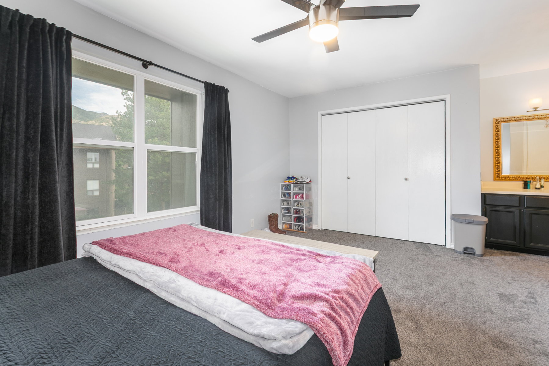 Bright bedroom featuring a large window, ceiling fan with integrated lighting, bi-fold closet doors, and built-in vanity with a gold-framed mirror