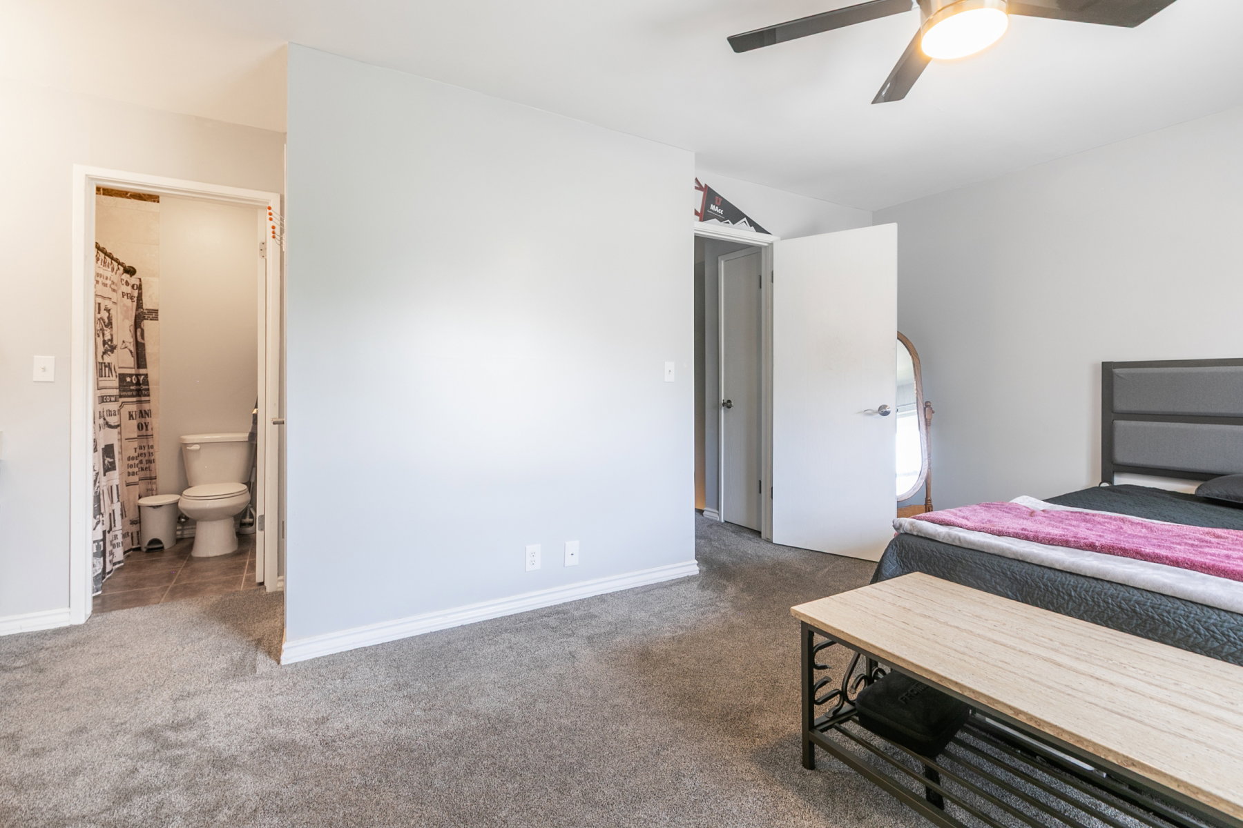 Carpeted room featuring a ceiling fan with integrated lighting, a solid-core door, and an entrance to a bathroom with tile flooring and a toilet