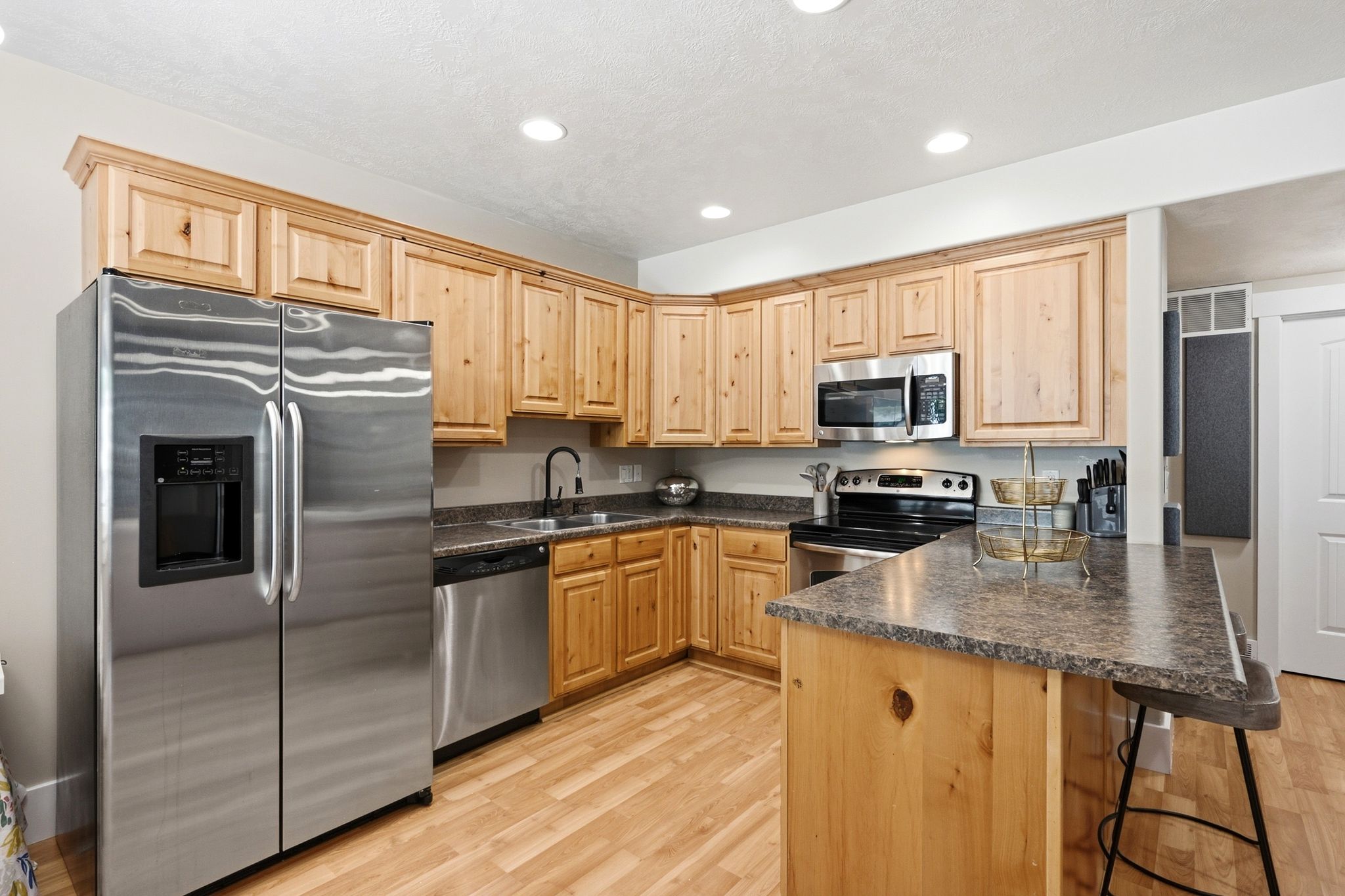 Kitchen featuring light wood cabinetry, stainless steel appliances, dark stone-finish countertops, and wood-finish flooring