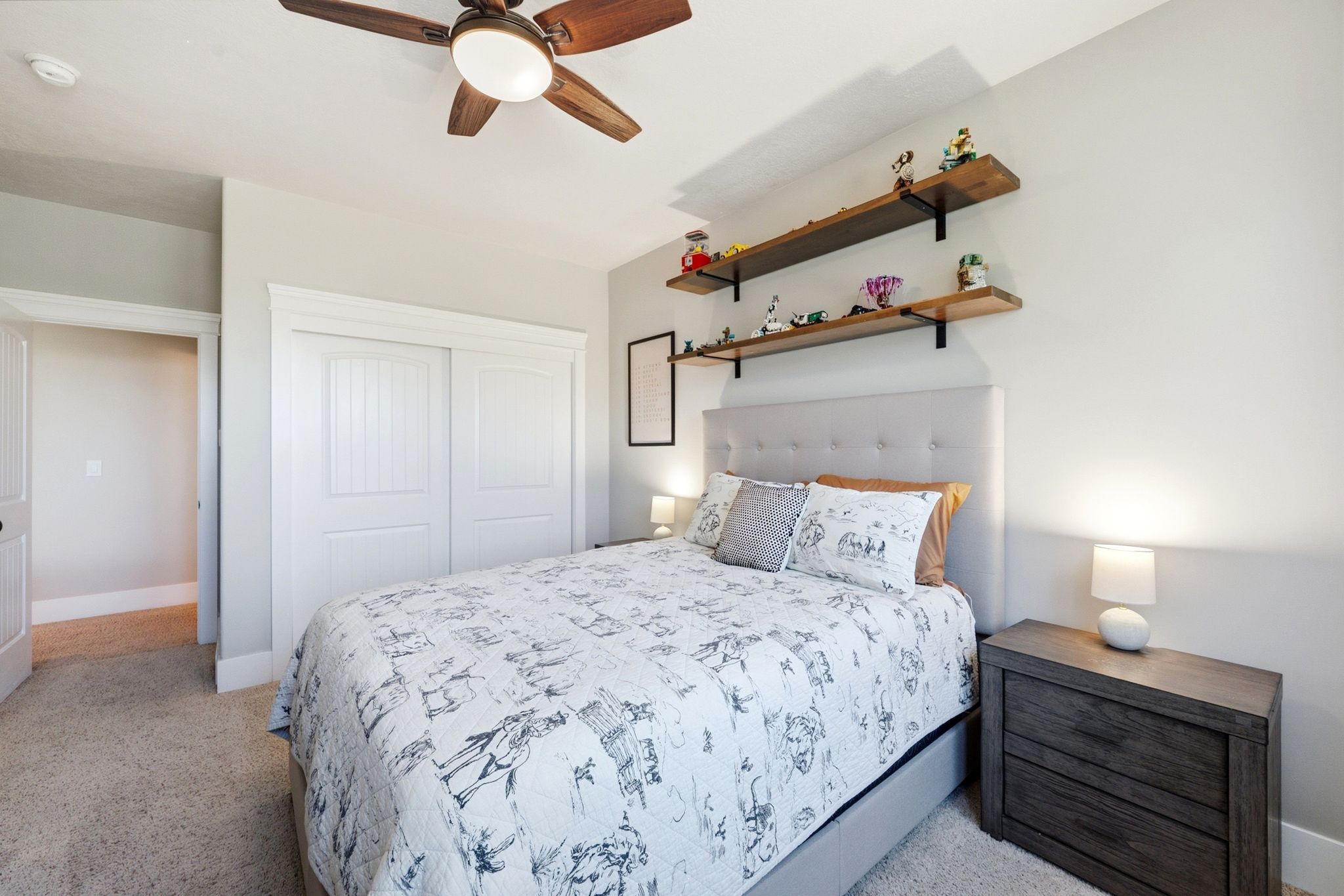 Bedroom featuring light-toned walls, carpeted flooring, a ceiling fan with light fixture, white sliding closet doors, and wood-finish floating shelves