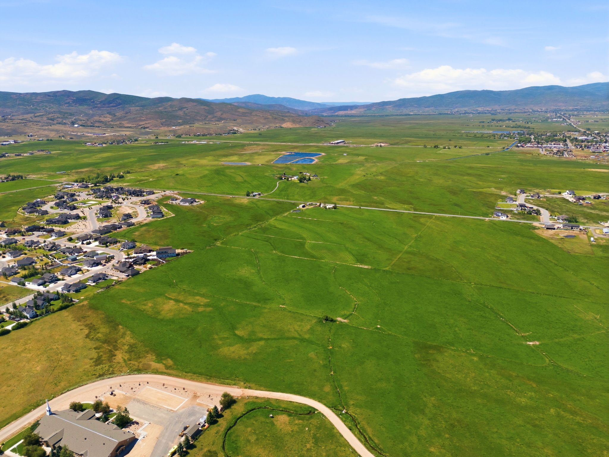 Expansive green landscape with distant mountains under a blue sky, featuring a residential development, open fields, and a body of water