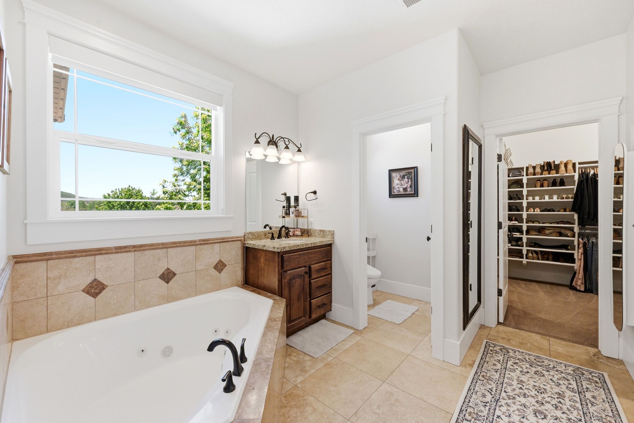 Soaking tub with tiled surround, dual-sink vanity with wood cabinetry and stone countertop, and separate water closet