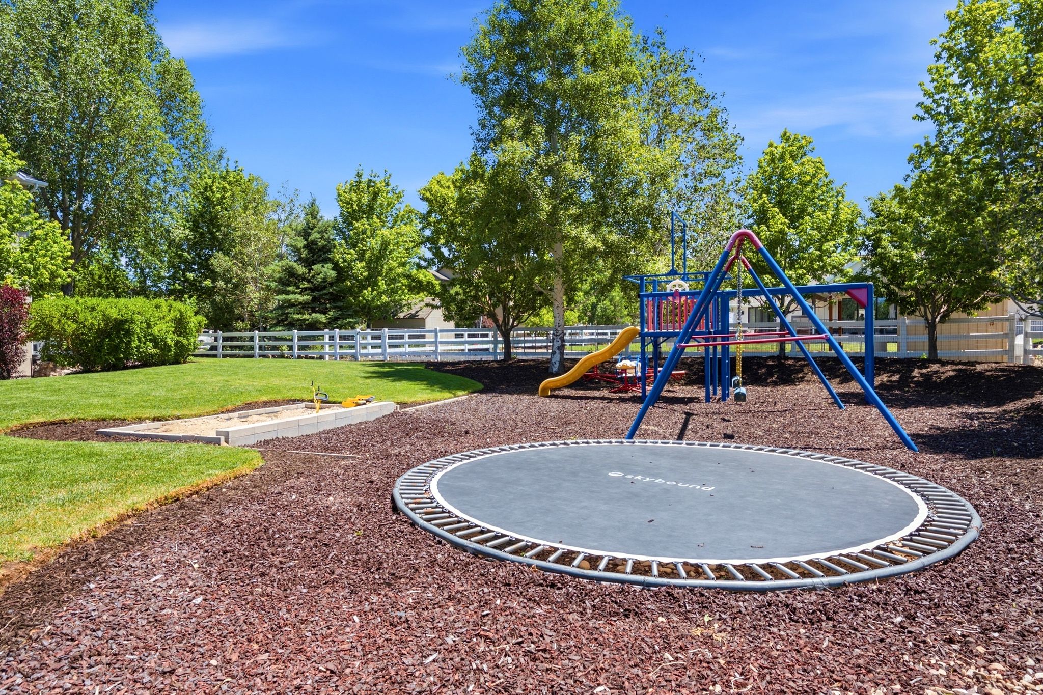Landscaped backyard featuring a built-in trampoline, a playset with a slide and swings, and a dedicated sandbox area