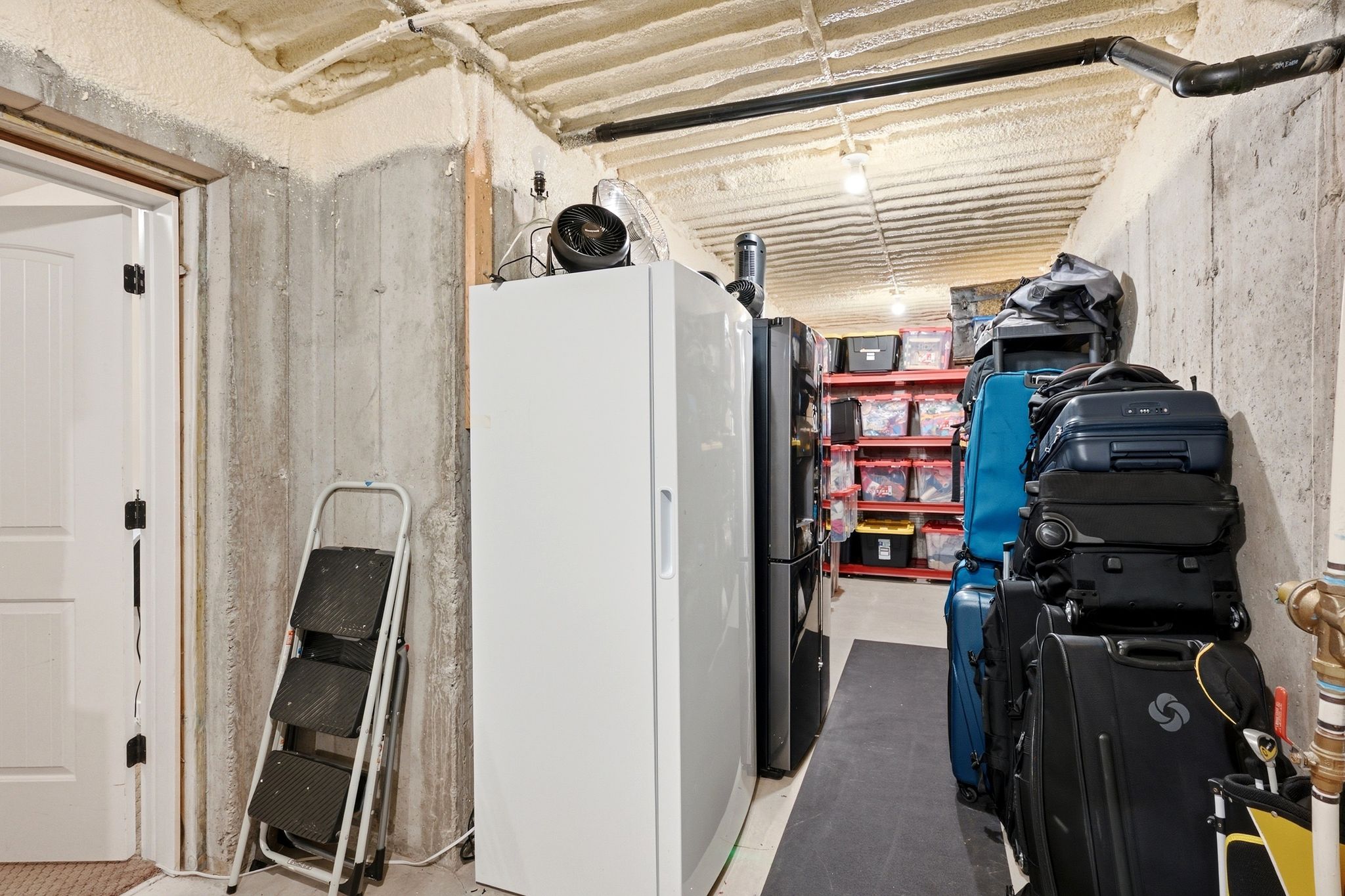 Unfinished basement space featuring exposed concrete walls, spray foam insulation on the ceiling, and overhead piping
