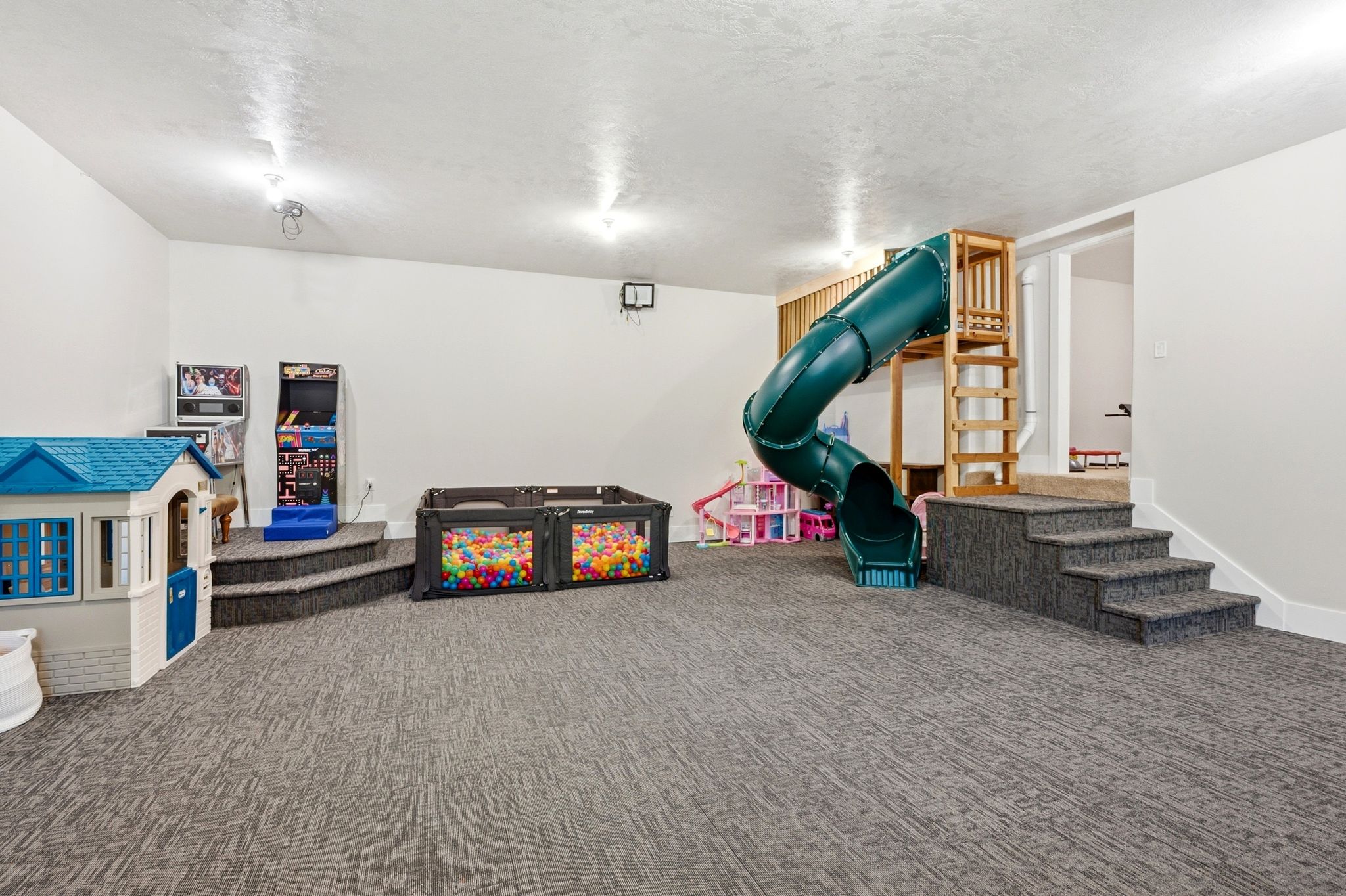 Spacious recreation room featuring a built-in play structure with a slide, recessed lighting, white walls, gray patterned carpet, and a raised platform