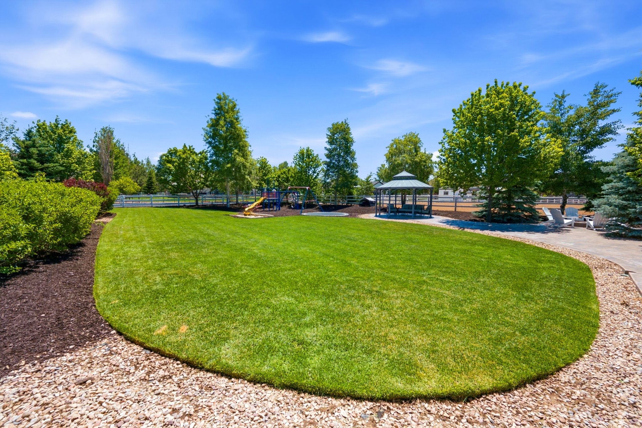 Expansive lawn area featuring a paved pathway, a built-in gazebo, mature trees, and a white perimeter fence