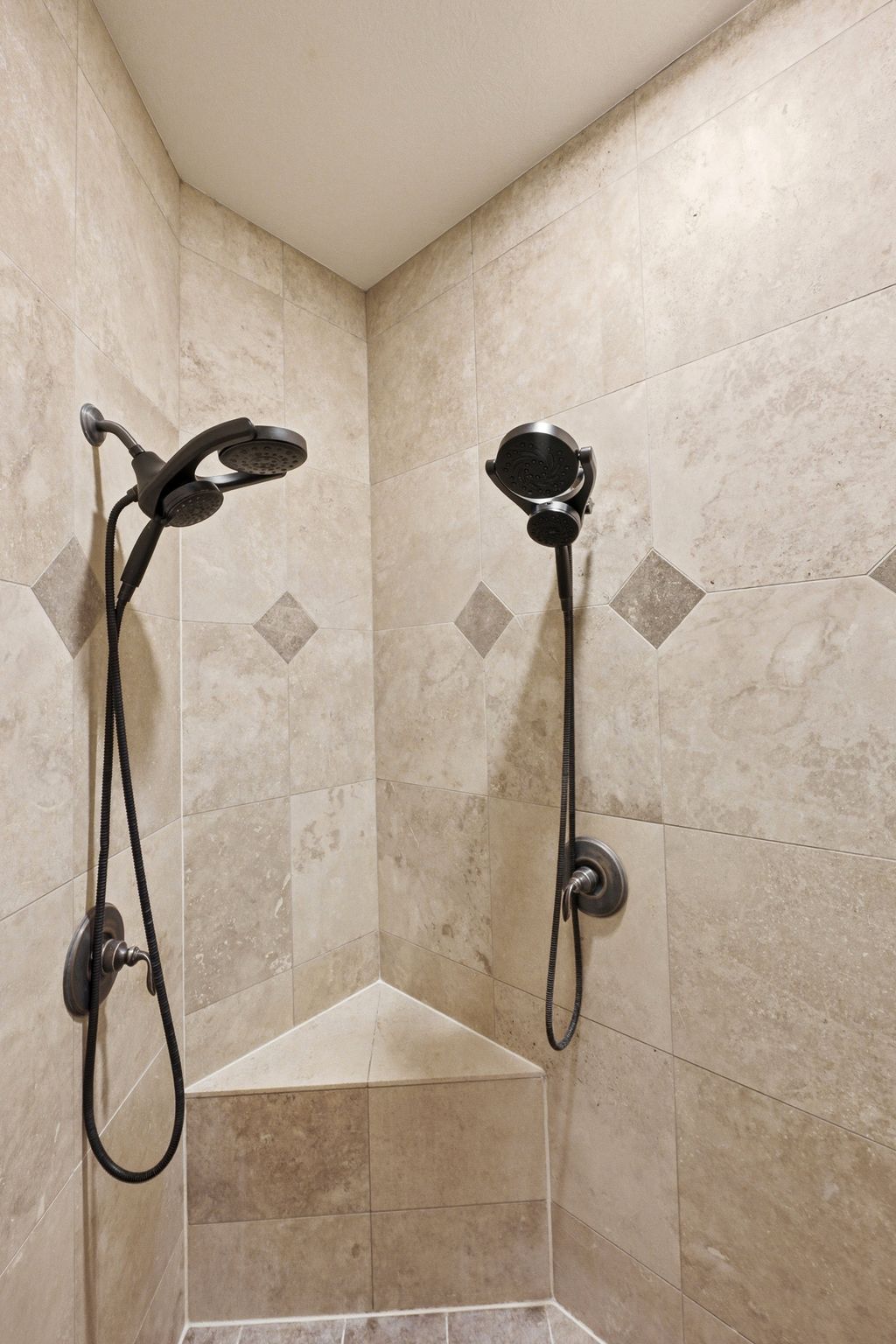 Tiled shower enclosure featuring dual shower heads with handheld sprayers, oil-rubbed bronze fixtures, an integrated corner bench, and a decorative tile accent band