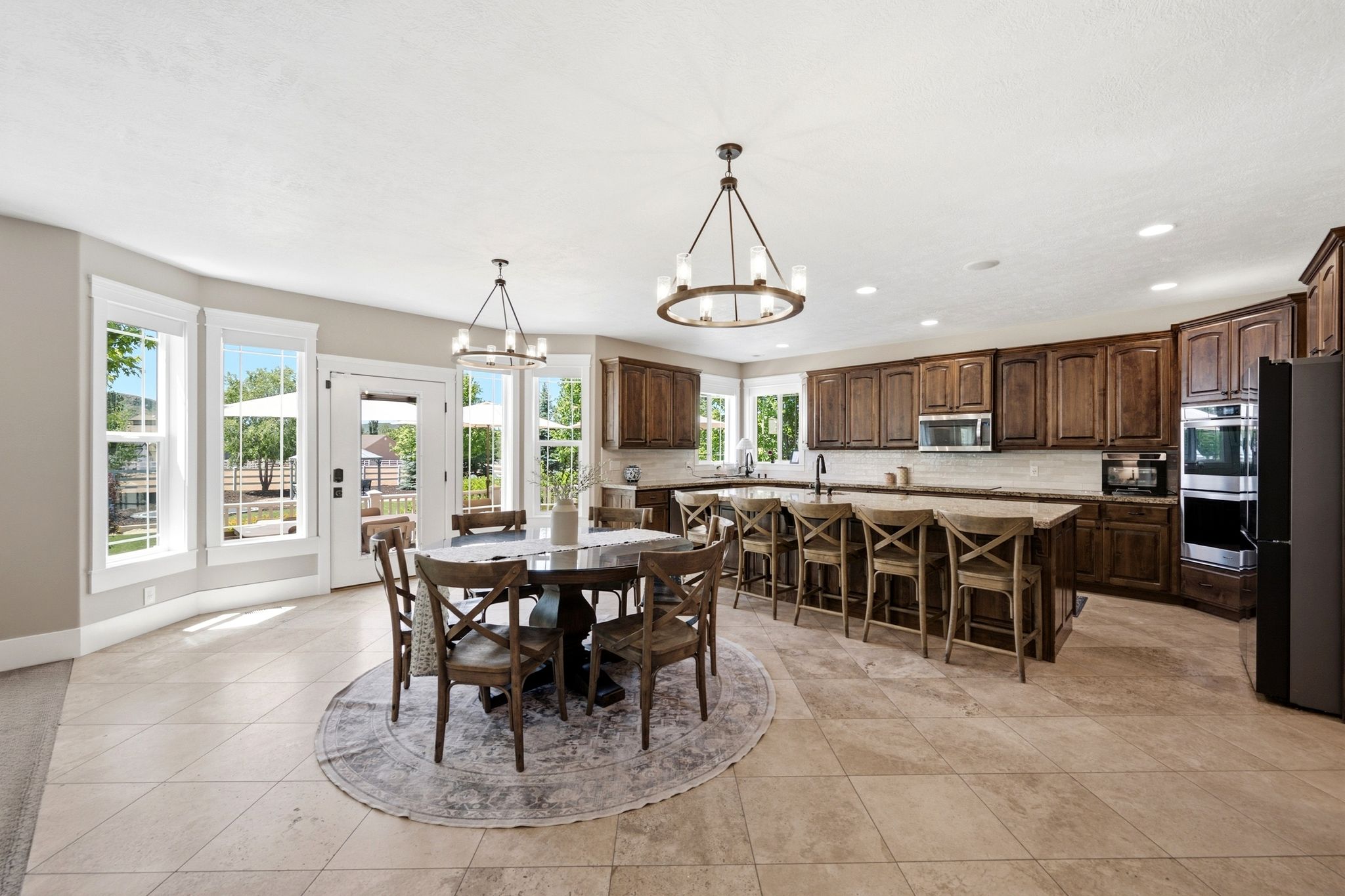 Spacious kitchen and dining area featuring stone tile flooring, dark wood cabinetry, a central island with seating, stainless steel appliances, and abundant natural light from multiple windows and a glass-paneled door