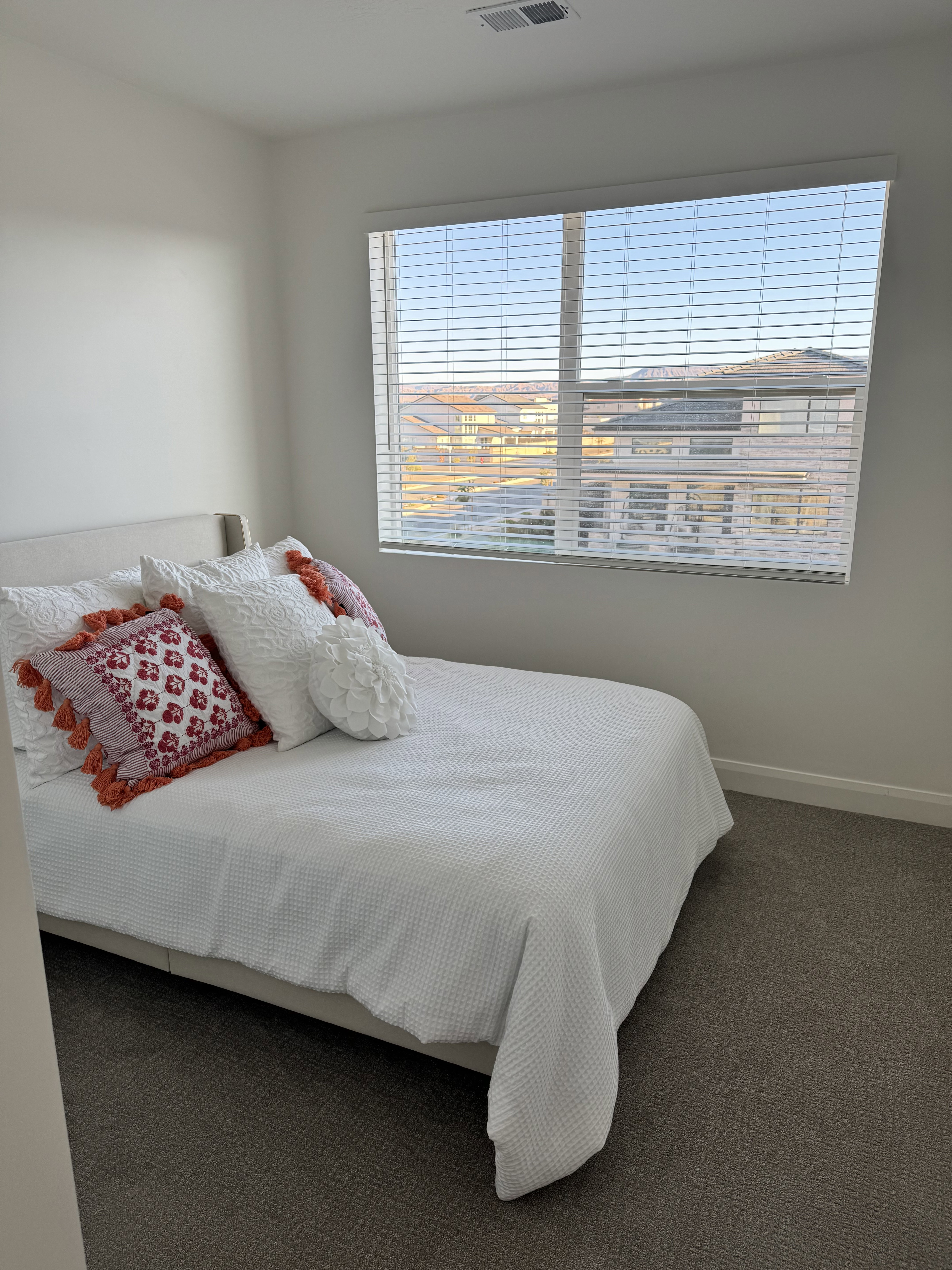 Carpeted room featuring a large window with horizontal blinds, offering distant views of residential rooftops
