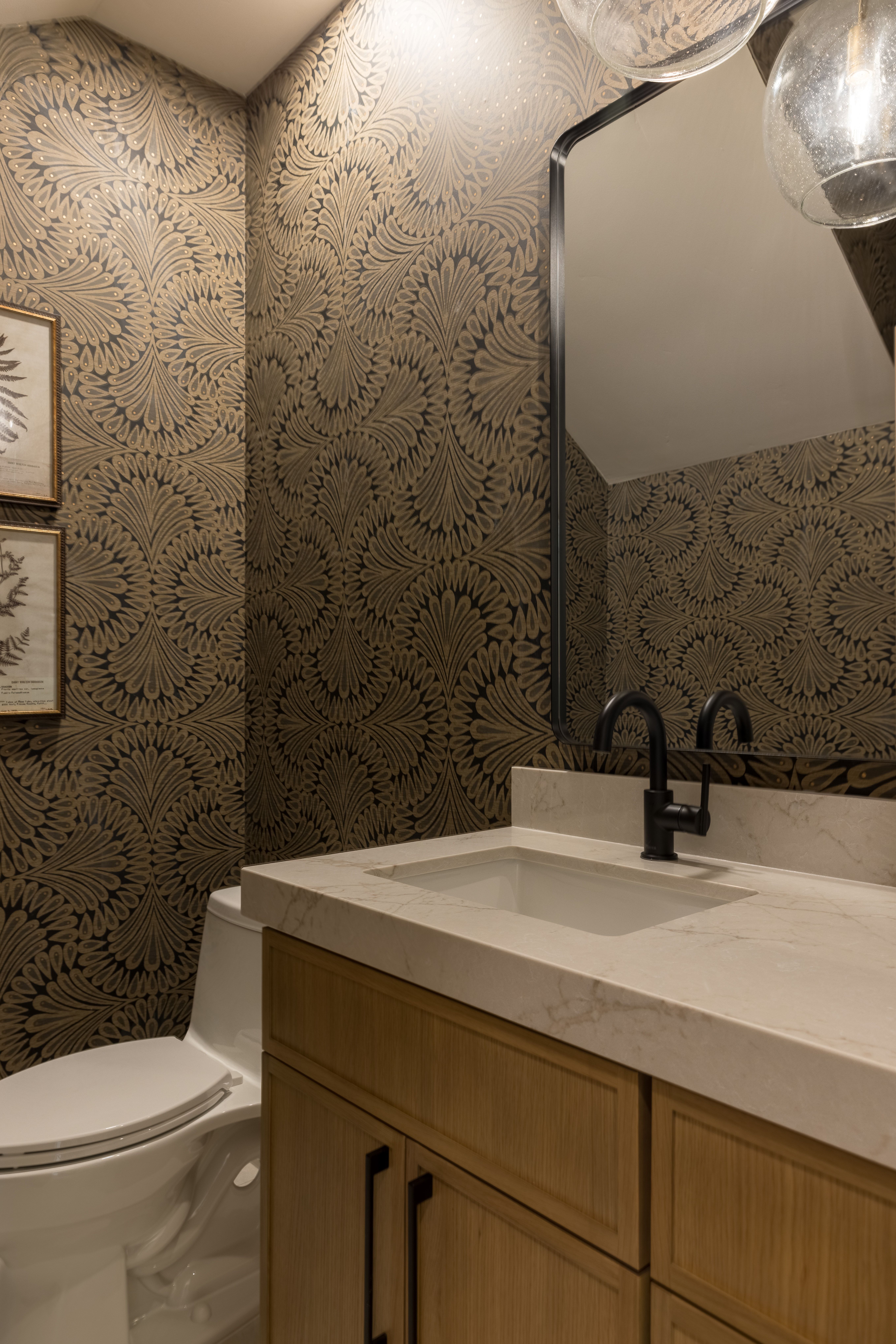 Powder room featuring intricate patterned wallpaper, a light wood-finish vanity with dark hardware, a white countertop with an integrated sink, a matte black faucet, and an arched mirror