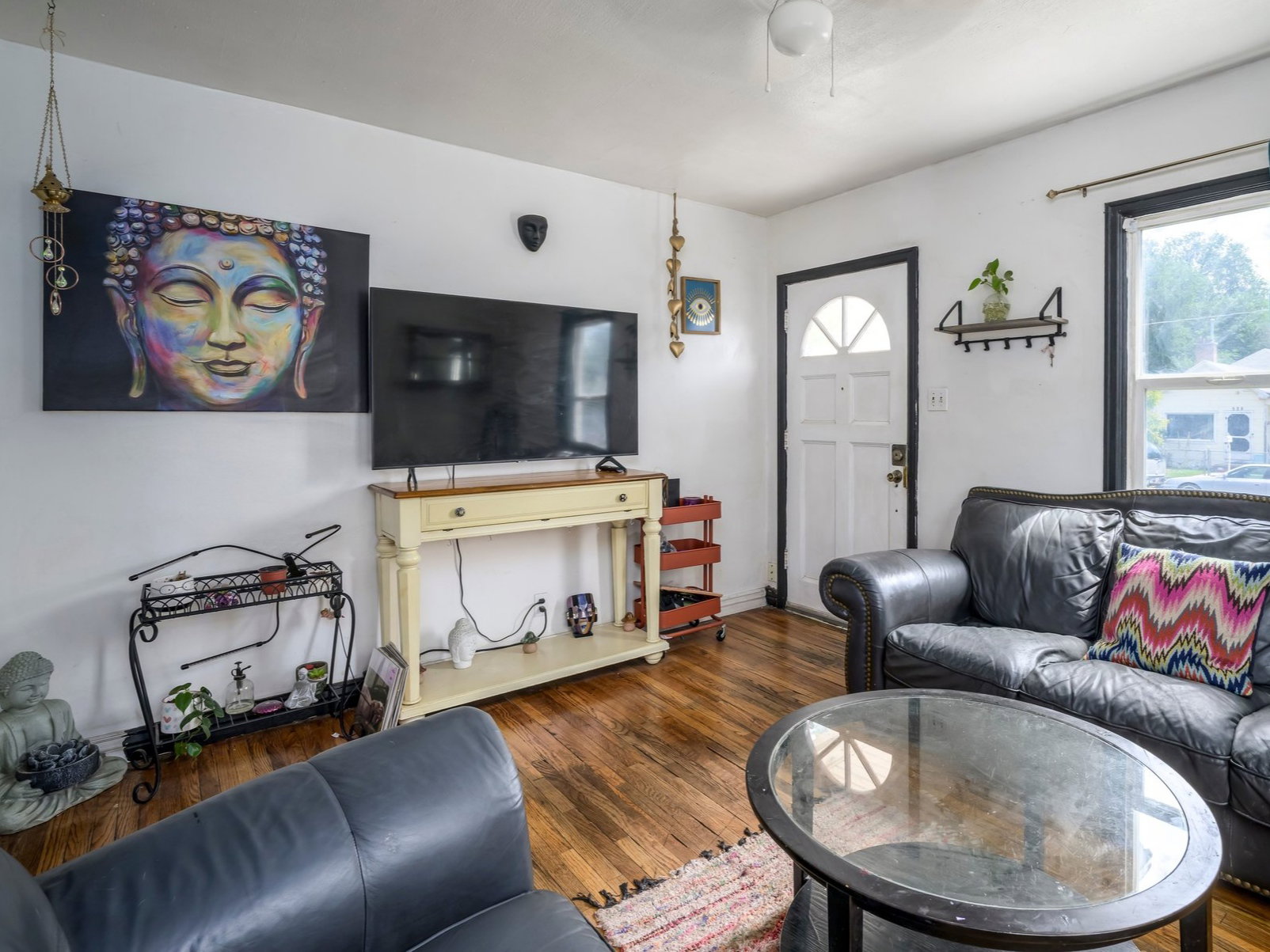 Spacious living area featuring warm wood-finish flooring, a white paneled entry door with an arched window, and a bright window with dark trim