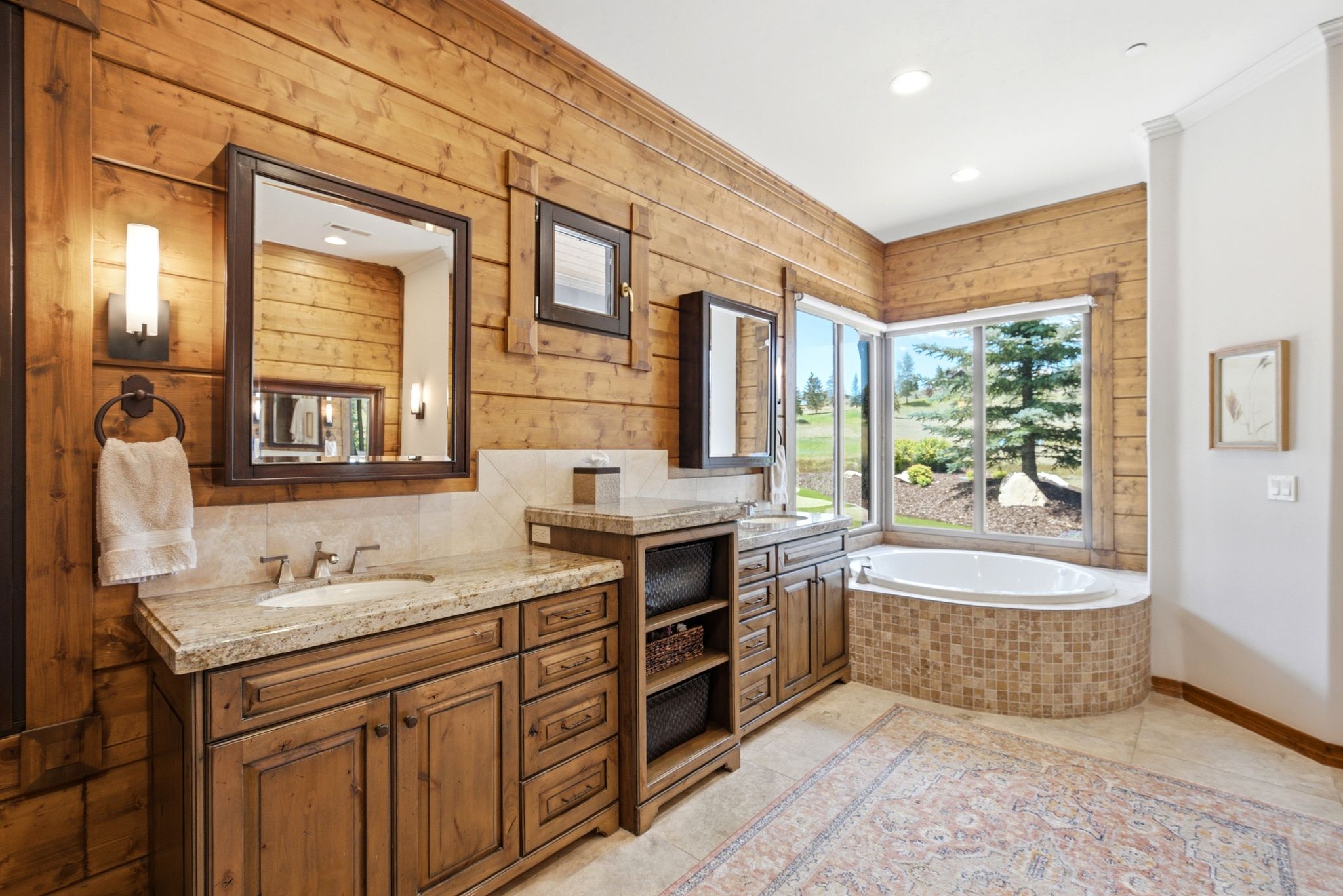 Expansive bath featuring a built-in soaking tub with mosaic tile surround, large window with outdoor landscaping, and dual wood-finish vanities with undermount sinks and light-toned countertops