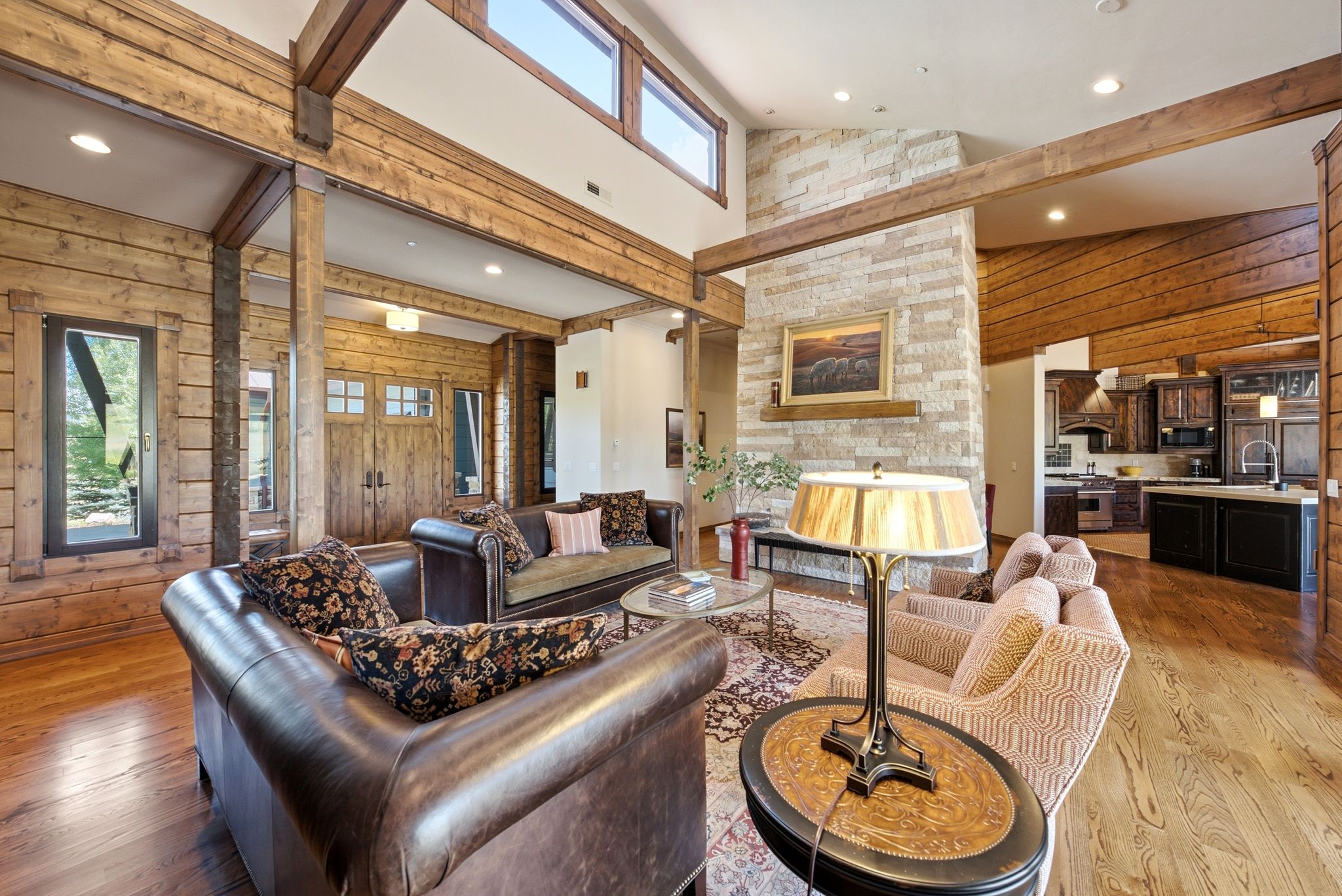 Great room featuring wood-finish flooring, exposed wood beams, a stone-clad fireplace with mantel, and a kitchen with dark cabinetry and an island