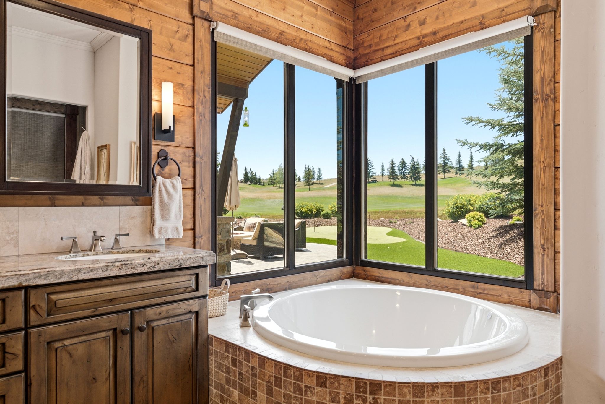 Built-in soaking tub with mosaic tile surround, corner windows, wood-finish paneling, vanity with stone countertop, and integrated sink