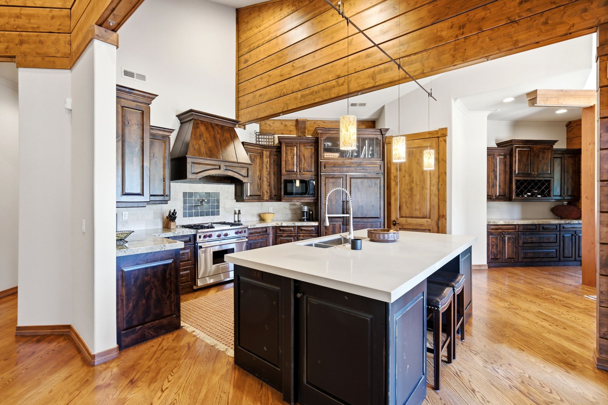 Gourmet kitchen featuring a large central island with light-toned countertops, rich wood-finish flooring, custom dark wood cabinetry, stainless steel appliances, and a custom wood range hood