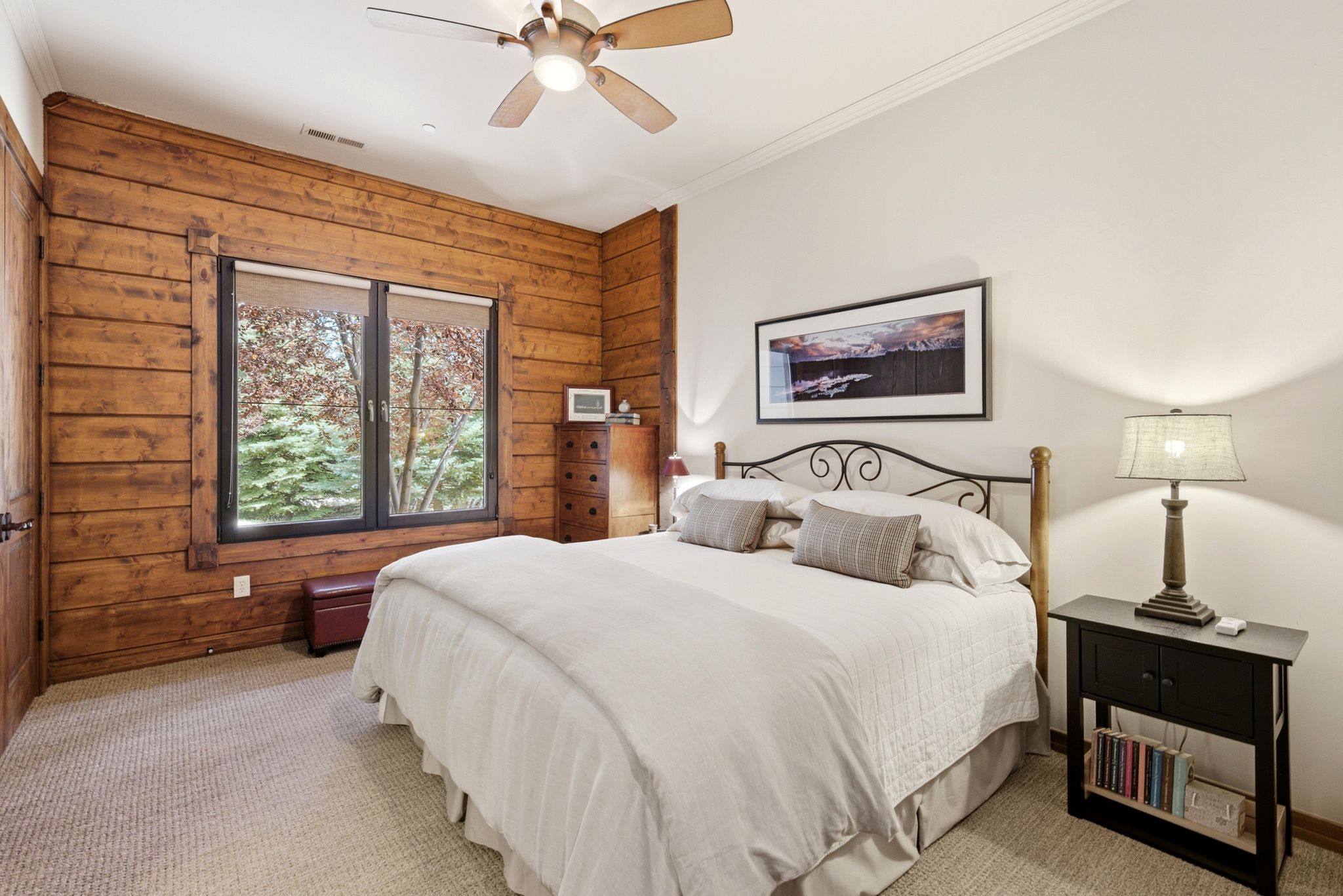 Sleeping area featuring a window with exterior scenery, wood-panel accent wall, textured carpet, and a ceiling fan with integrated lighting