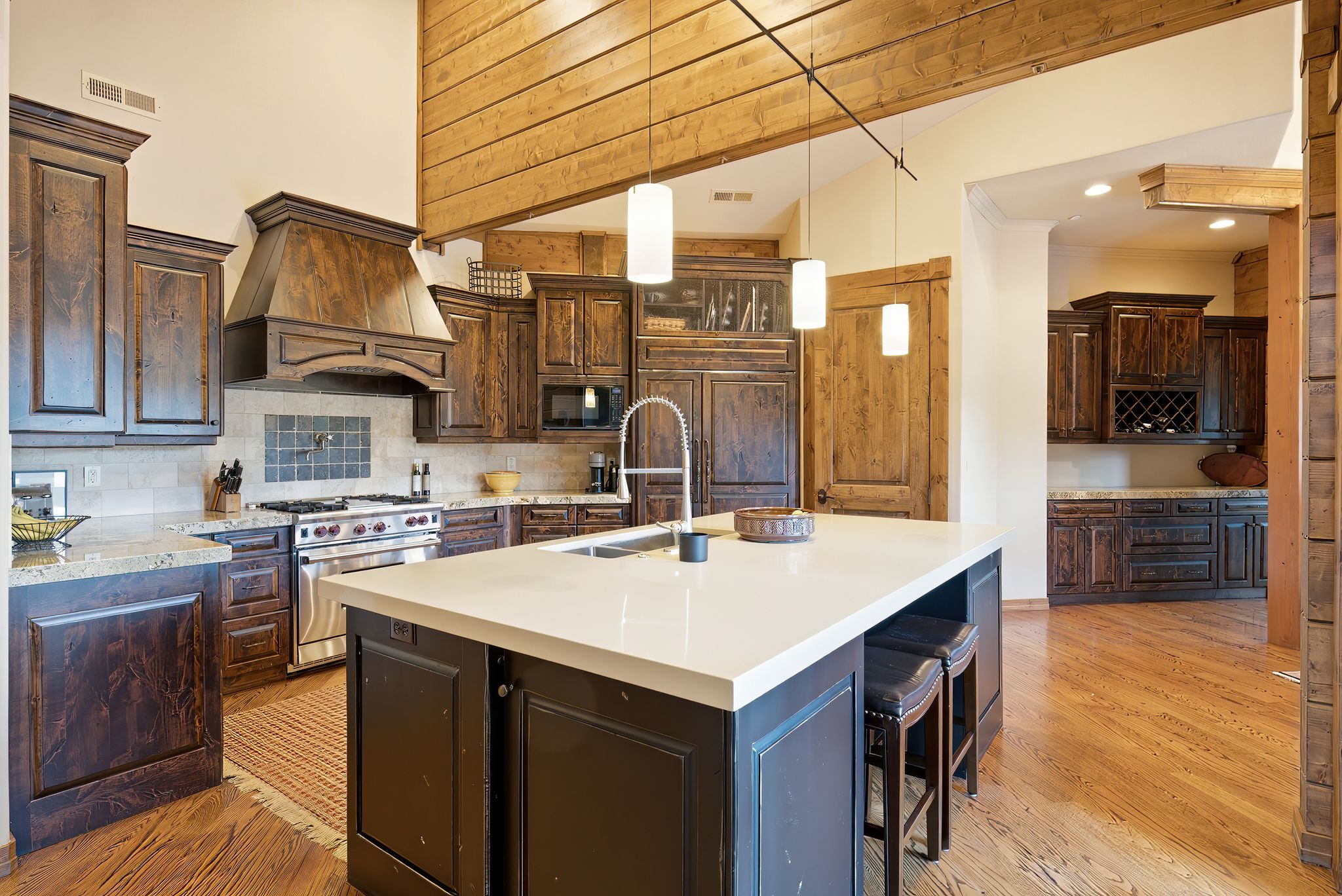 Gourmet kitchen featuring a large central island with a solid surface countertop, stainless steel range with custom wood hood, dark wood cabinetry, light wood-finish flooring, and recessed and pendant lighting