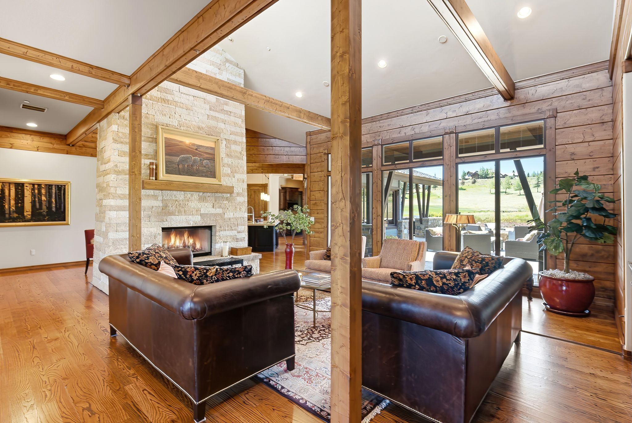 Living area featuring a stone-clad fireplace with a hearth, exposed wood beams, wood-finish flooring, wood plank accent walls, and expansive windows