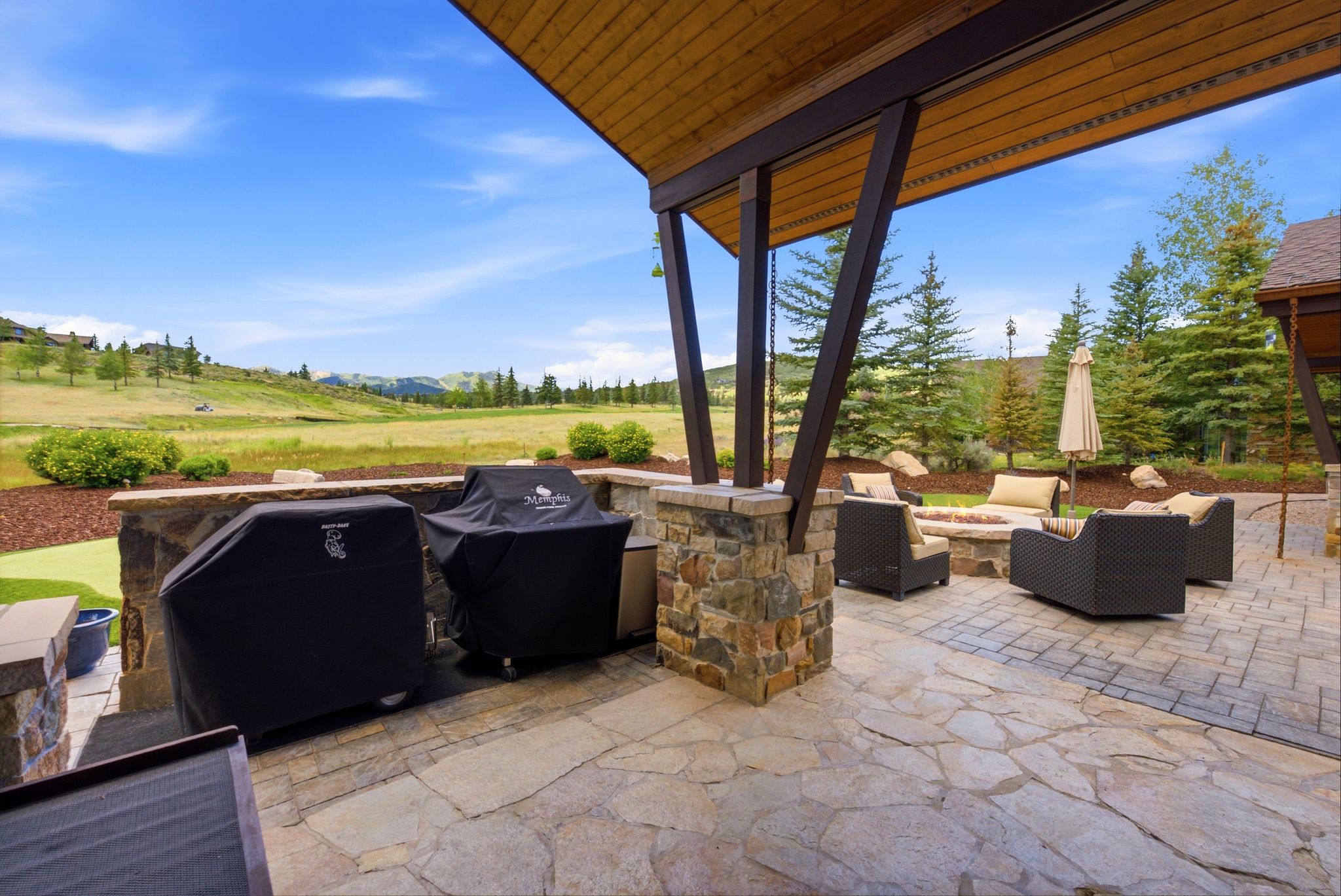 Expansive flagstone patio featuring a stone-clad outdoor kitchen area, covered outdoor living space with wood ceiling, and a built-in stone fire pit
