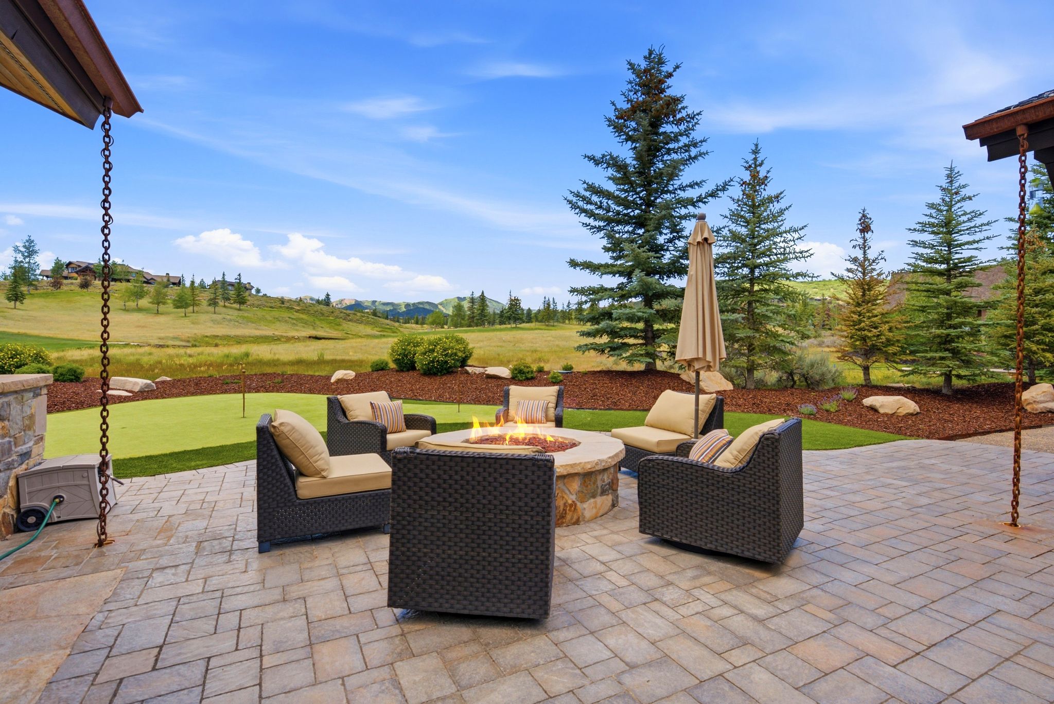 Spacious paver patio with a built-in stone fire pit, a putting green, and mature evergreen trees