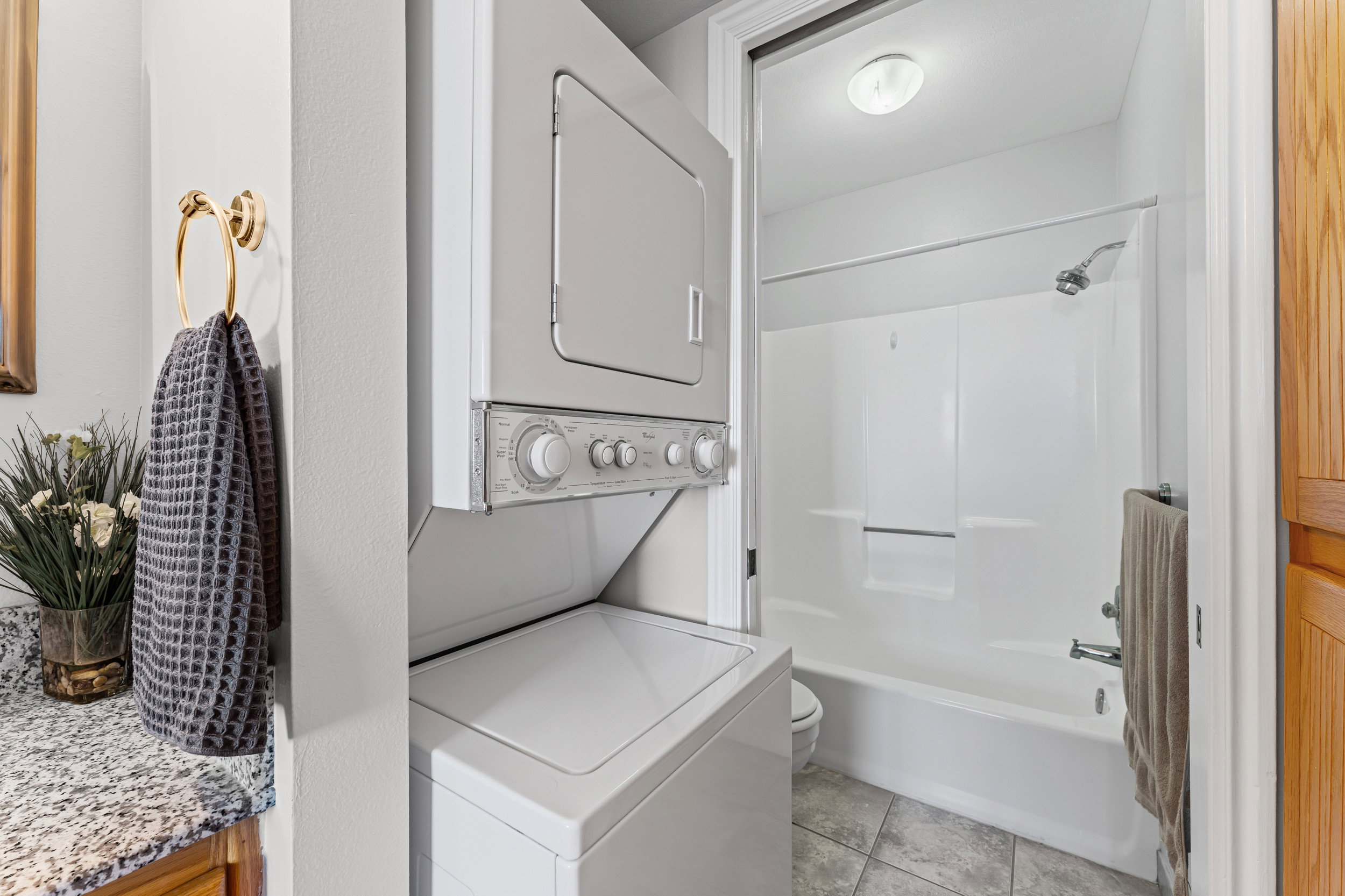 Compact laundry area with stackable washer and dryer, adjacent to a full bathroom featuring a white tub/shower combination, tile flooring, and a ceiling-mounted light fixture