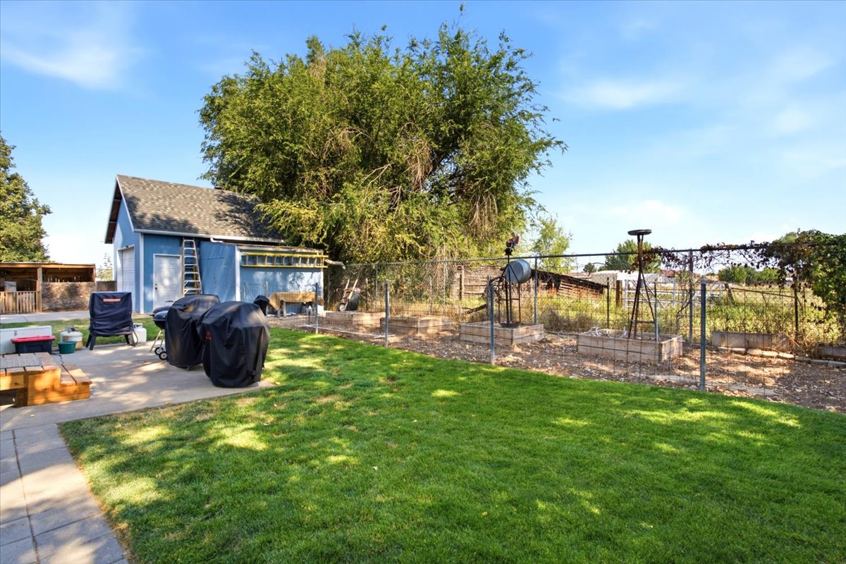 Expansive backyard featuring a manicured lawn, concrete patio, a detached utility building with a shingle roof, established trees, and fenced garden beds