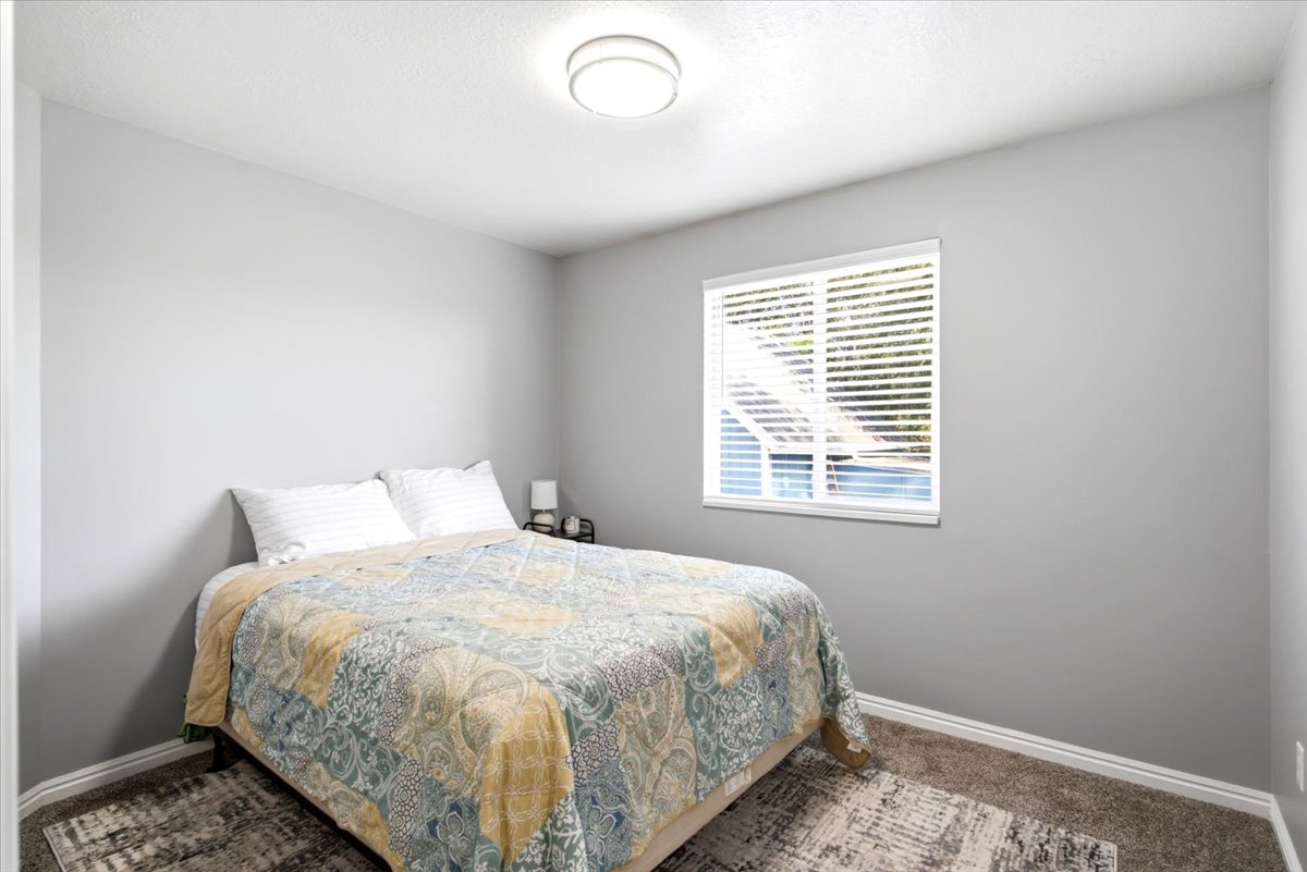 Bedroom featuring light gray wall paint, a single window with horizontal blinds, textured carpet flooring, and white trim work