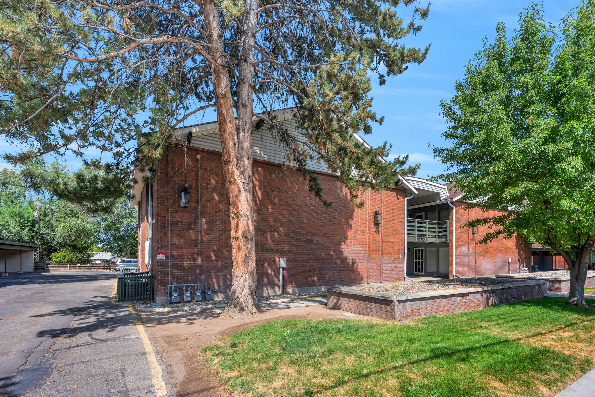 Brick exterior building featuring a covered entrance with a white railing balcony above, mature trees, and a grass lawn
