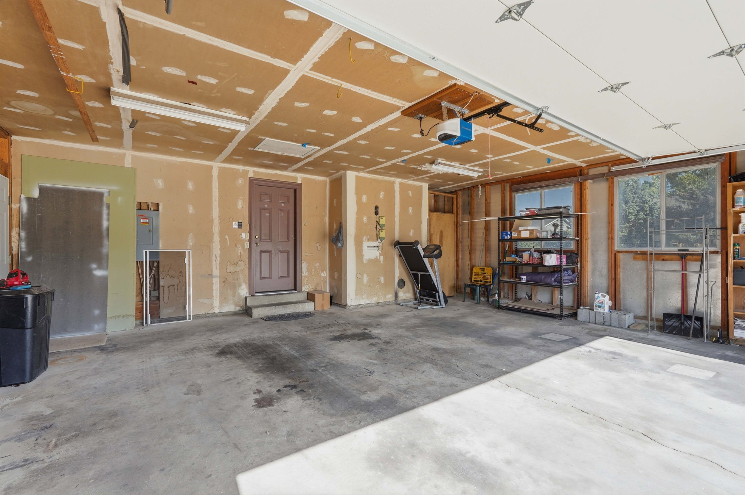 Spacious garage featuring a concrete floor, exposed drywall and framing, a brown door with steps, and multiple windows providing natural light