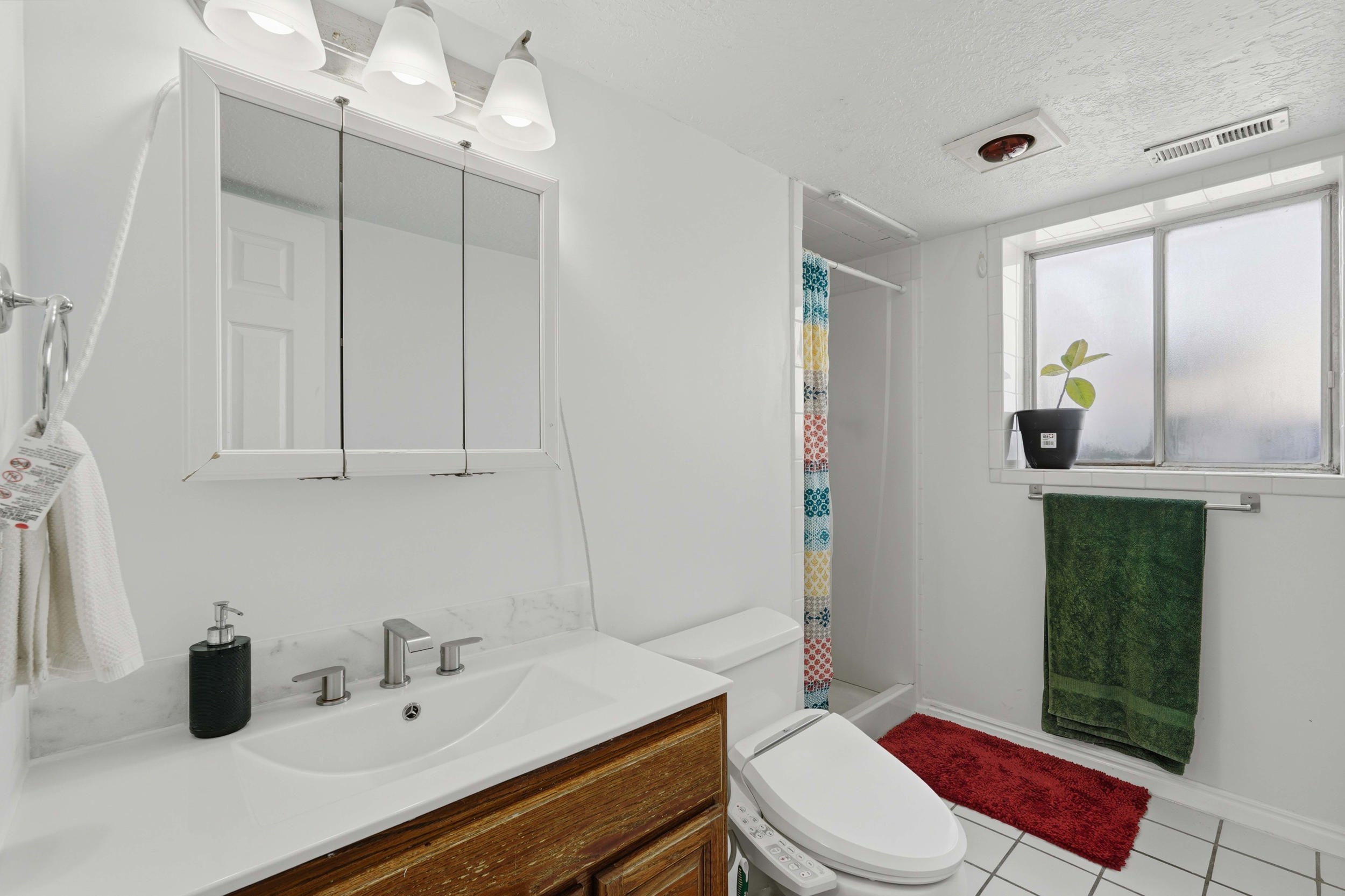 Bathroom featuring a wood-finish vanity with a white countertop and integrated sink, a three-panel mirrored medicine cabinet, a shower stall, a window, and white square floor tiles