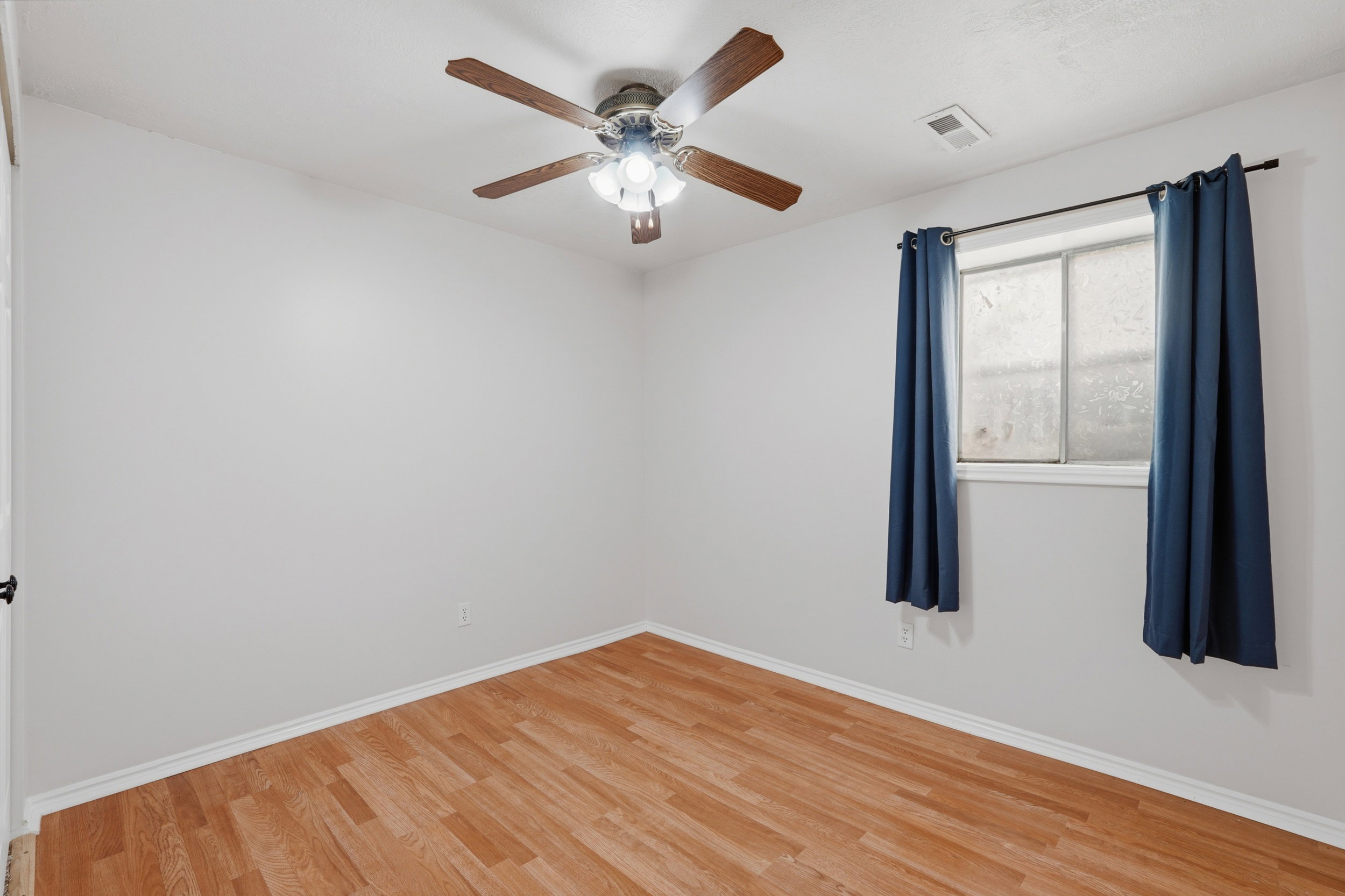 Bright interior space featuring wood-finish flooring, a ceiling fan with integrated lighting, and a window with blue curtain panels