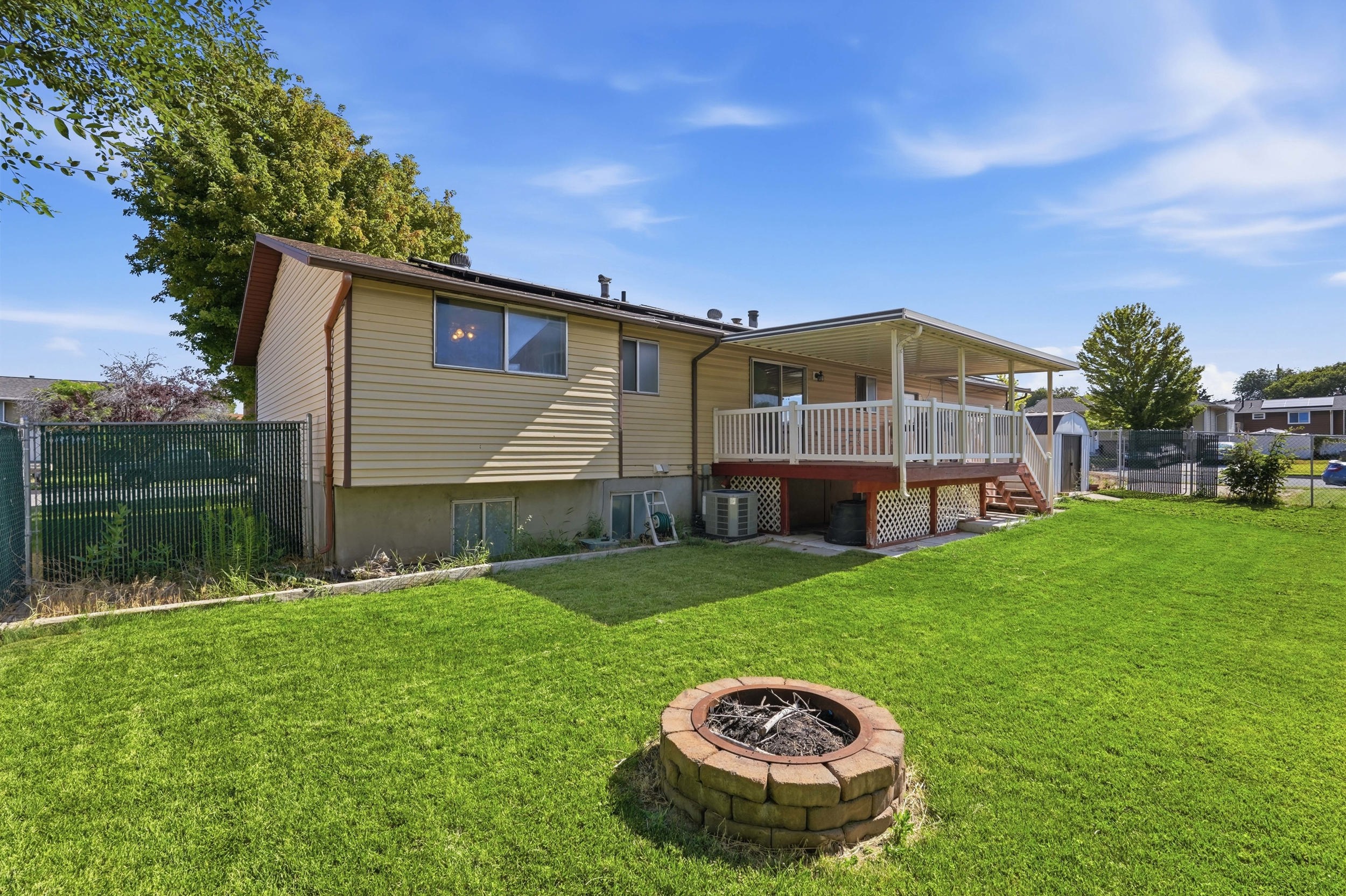 Expansive backyard featuring a covered deck, wood deck railing, a brick fire pit, and a chain-link fence