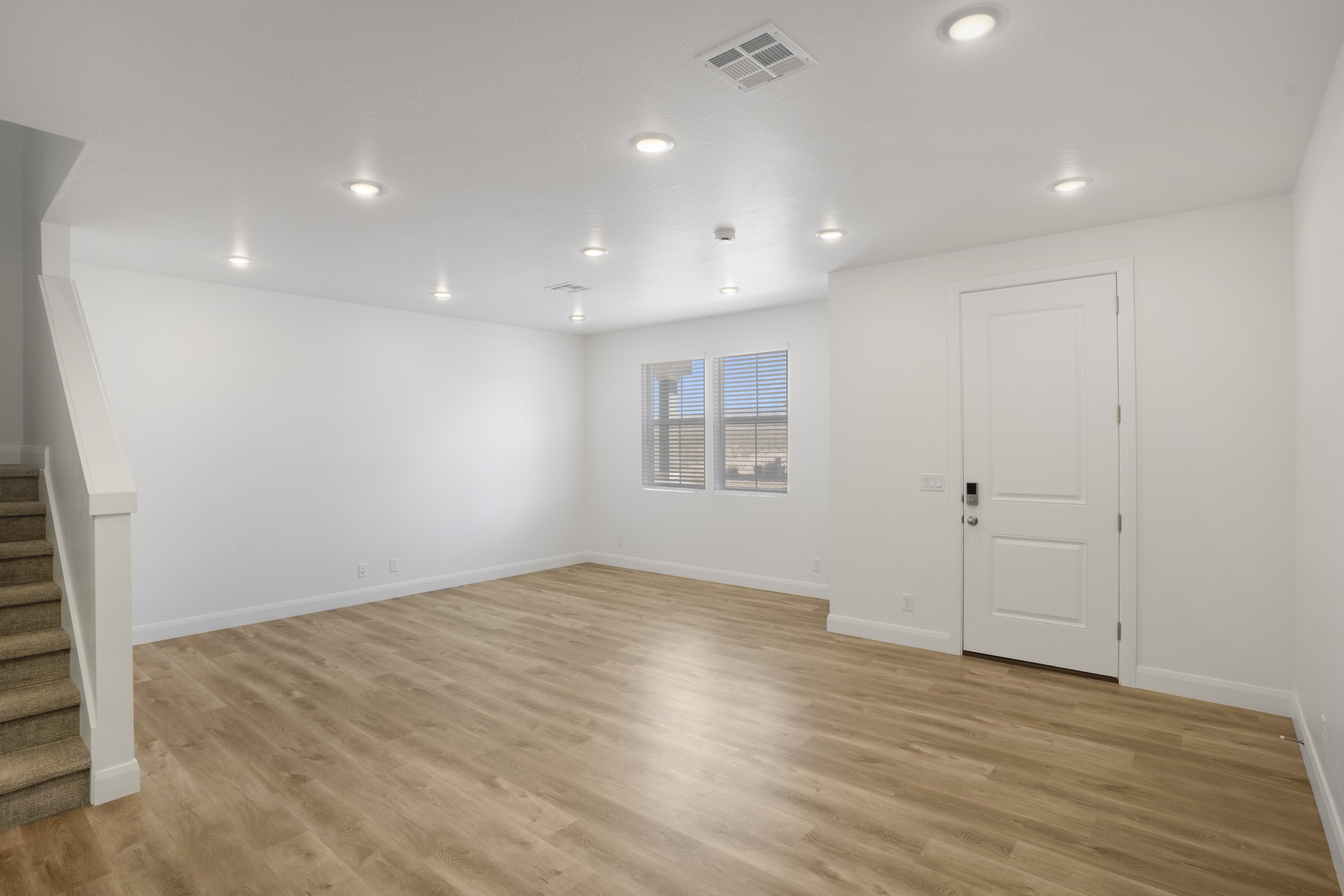 Spacious room featuring wood-finish flooring, recessed ceiling lighting, a white panel door with modern hardware, and multiple windows with blinds