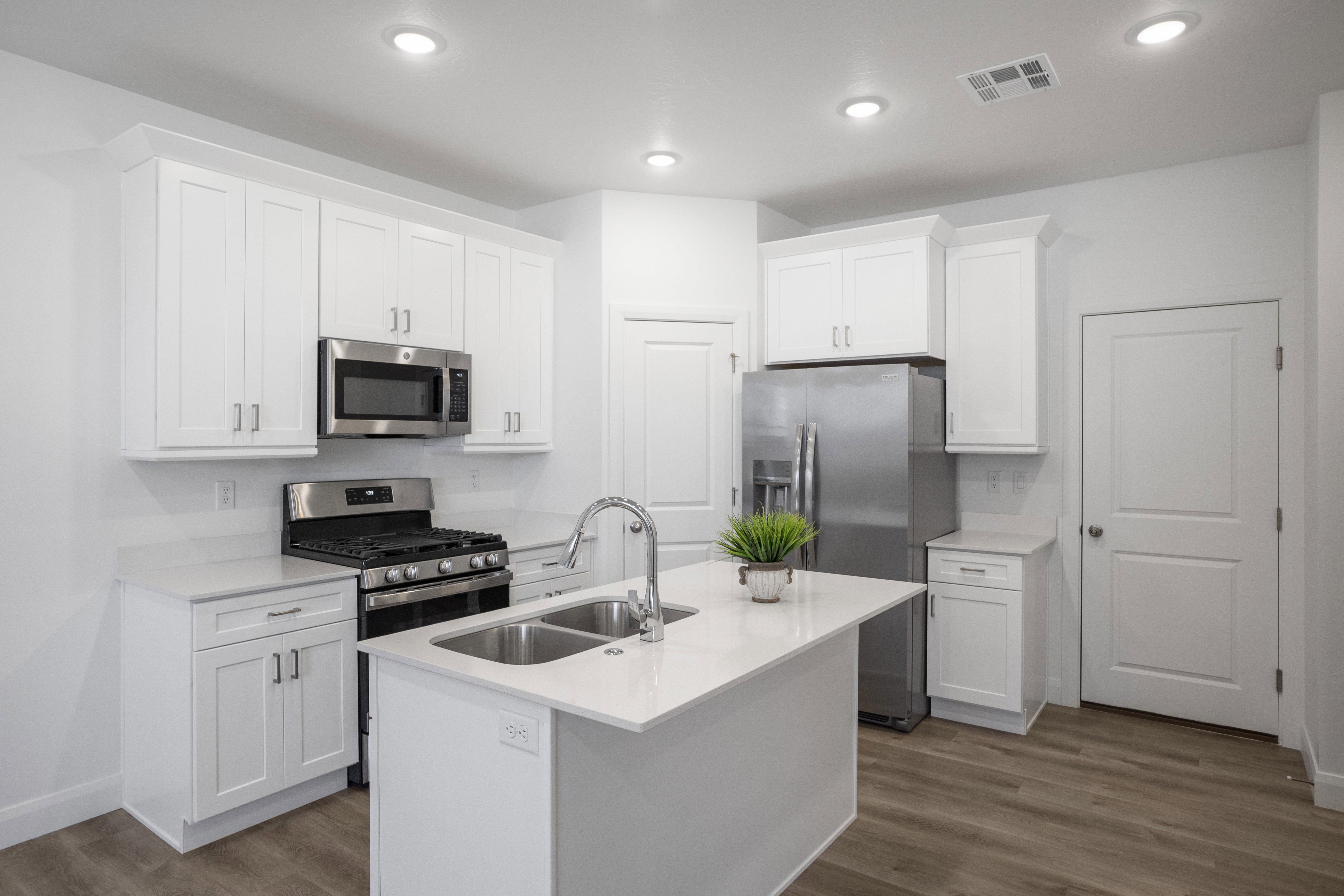 Contemporary kitchen featuring white shaker cabinetry, stainless steel appliances, a center island with a double basin sink, light-toned countertops, and wood-finish flooring