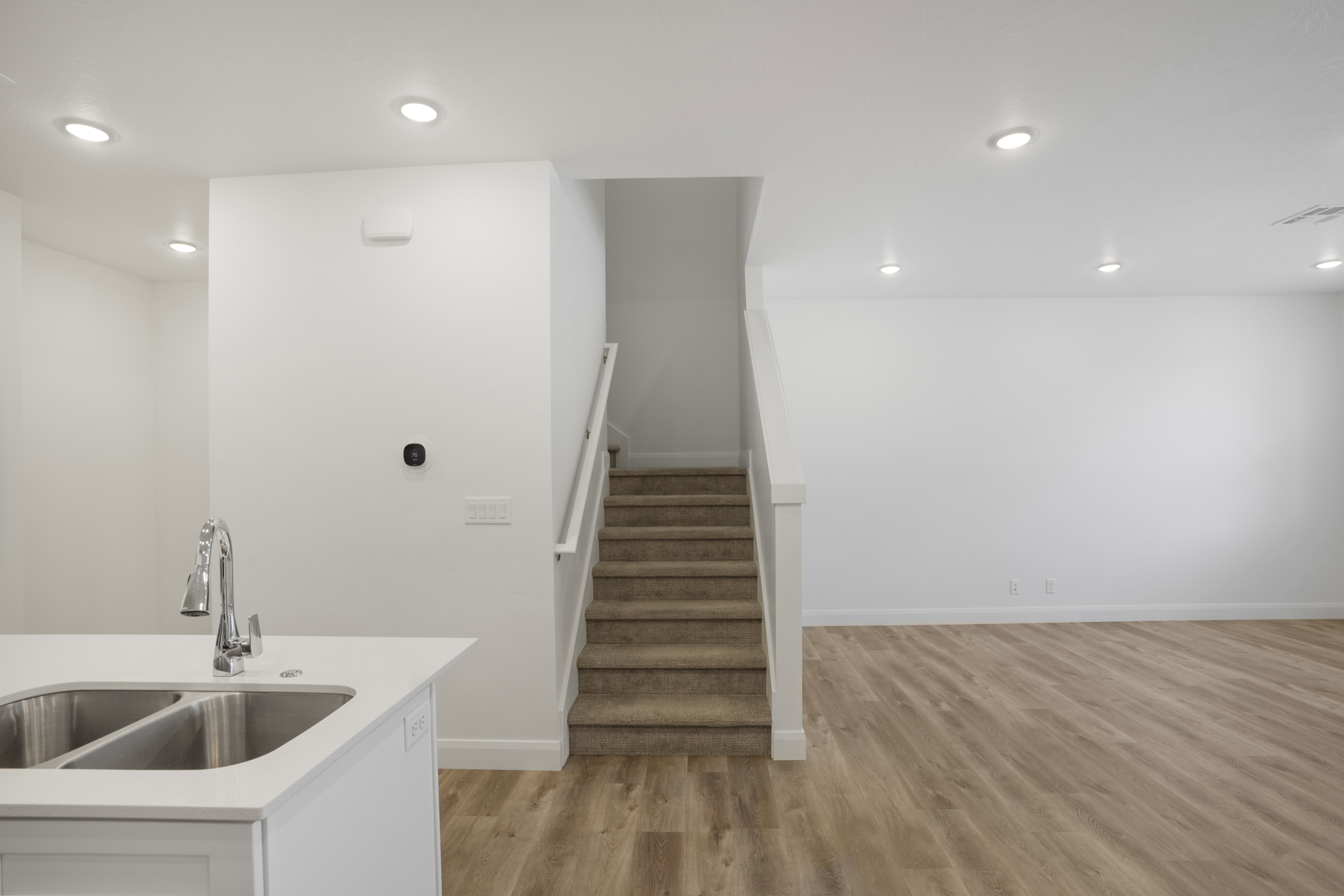 Open-concept living space featuring wood-finish flooring, recessed lighting, a white painted staircase with carpeted treads, and a kitchen island with a double stainless steel sink and a chrome pull-down faucet
