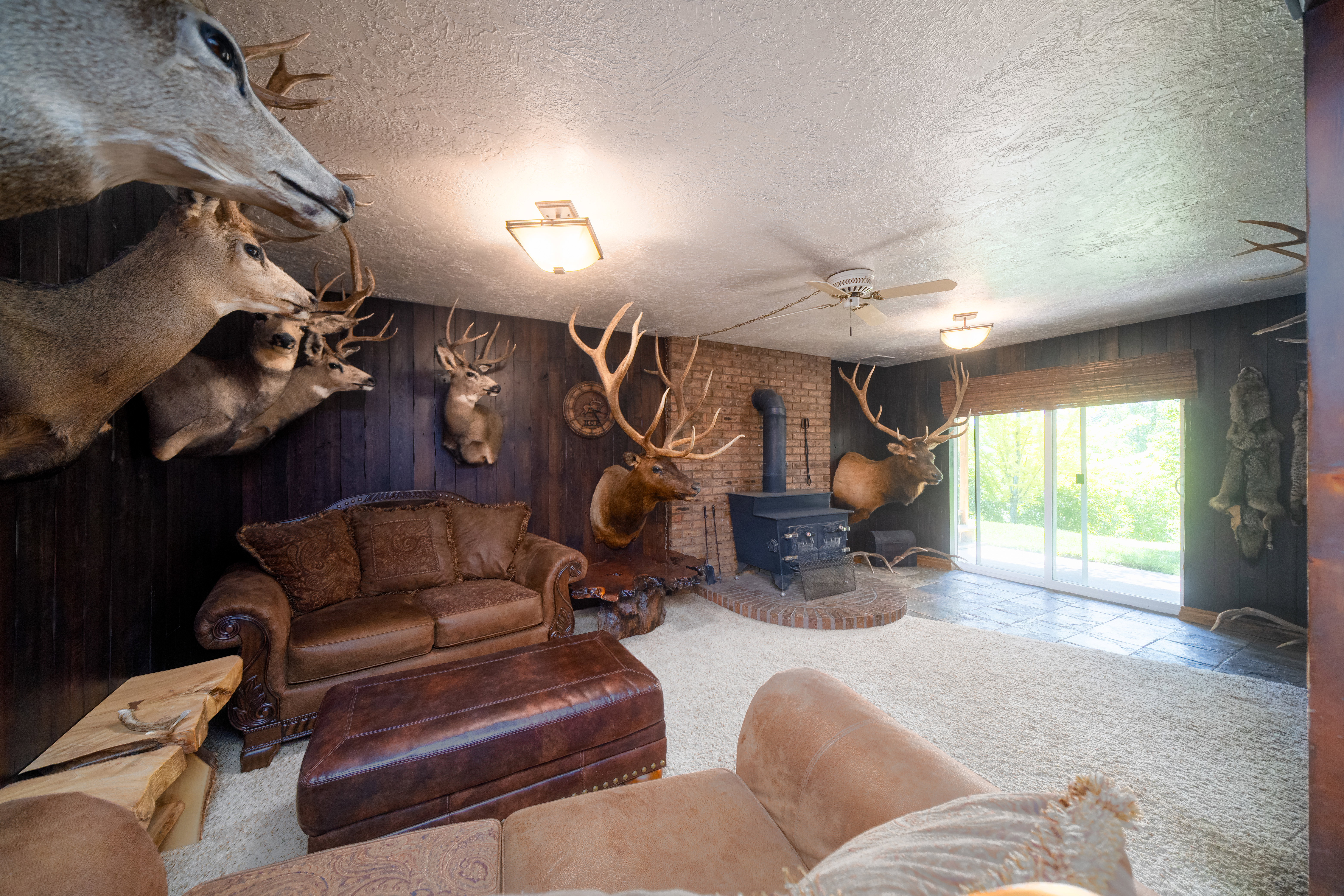 Spacious room featuring dark wood-paneled walls, a brick accent wall with a wood-burning stove, a ceiling fan, sliding glass doors, and light-colored carpeting