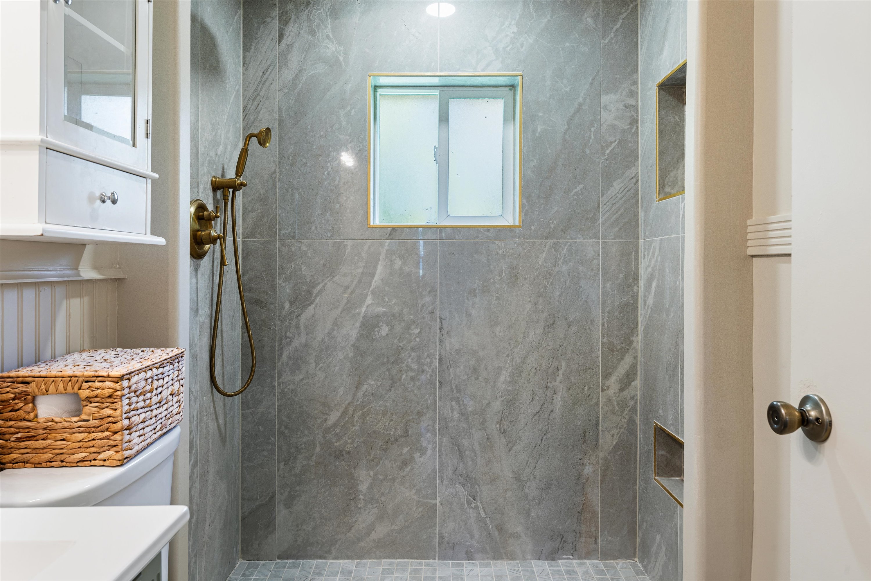 Shower featuring large format gray stone-finish wall tile, a frosted window with gold-tone trim, brass-finish shower fixtures, and a tiled shower niche with gold-tone trim