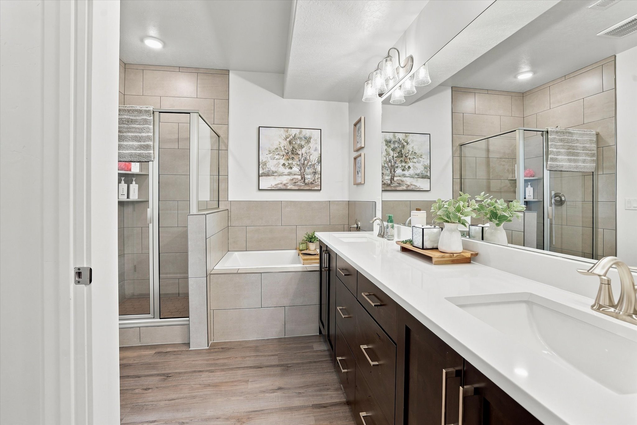 Contemporary bathroom featuring a double vanity with ample storage, a built-in soaking tub, a glass-enclosed shower, wood-finish flooring, and large format wall tiling