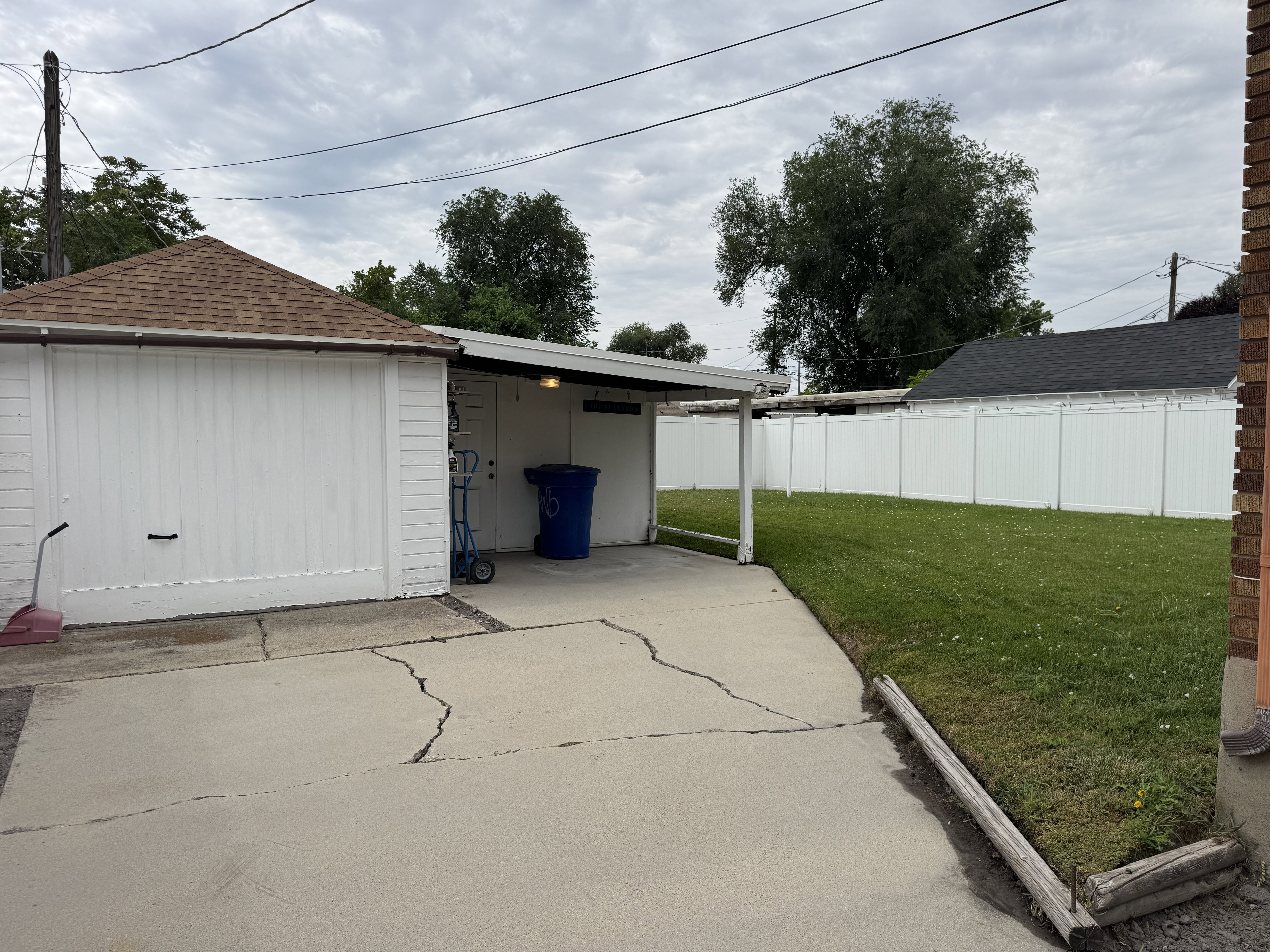 Private backyard space featuring a white vinyl privacy fence, a garage with a brown shingle roof, a covered carport, and a concrete patio area