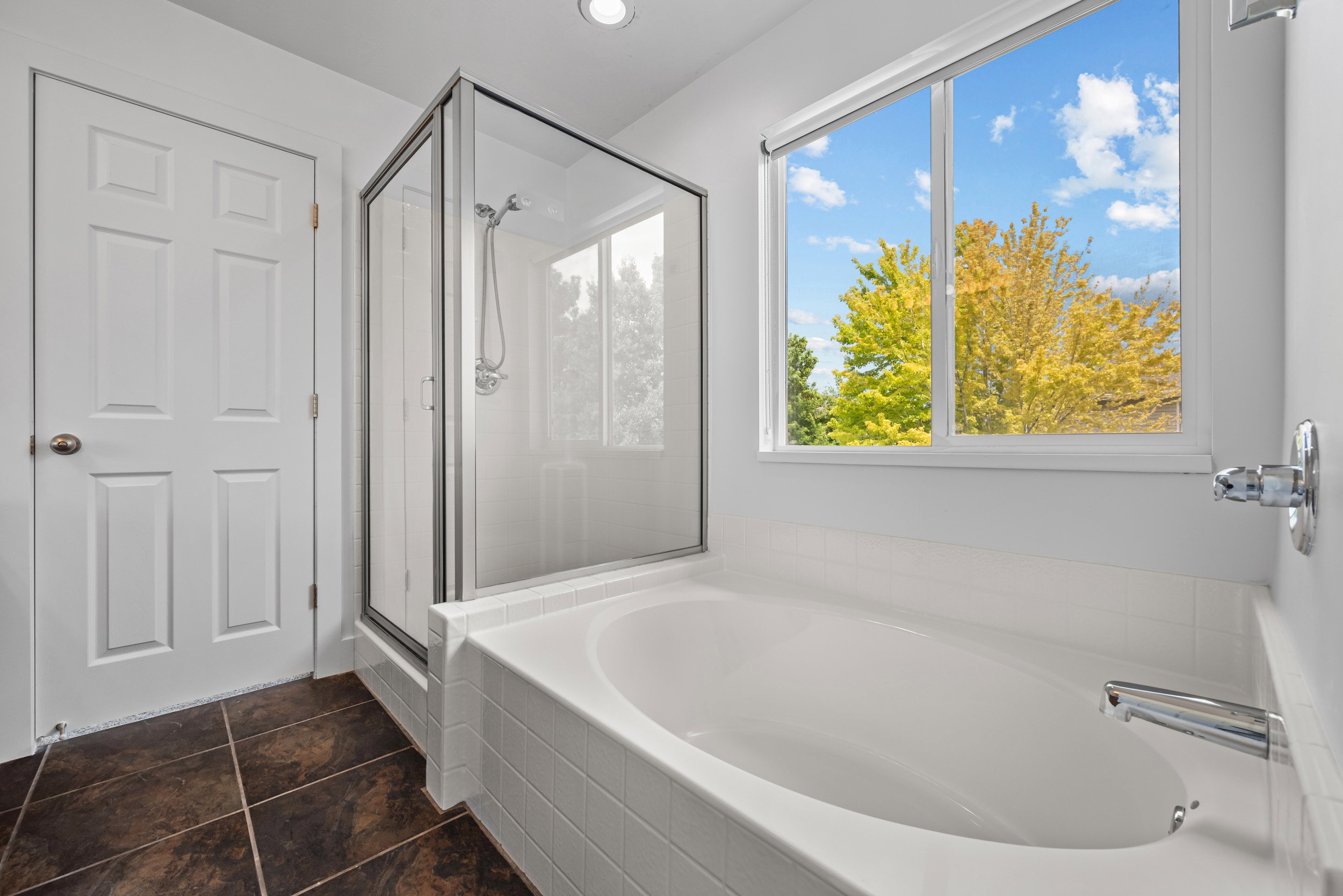 Soaking tub with white tile surround, glass-enclosed shower, and a large window